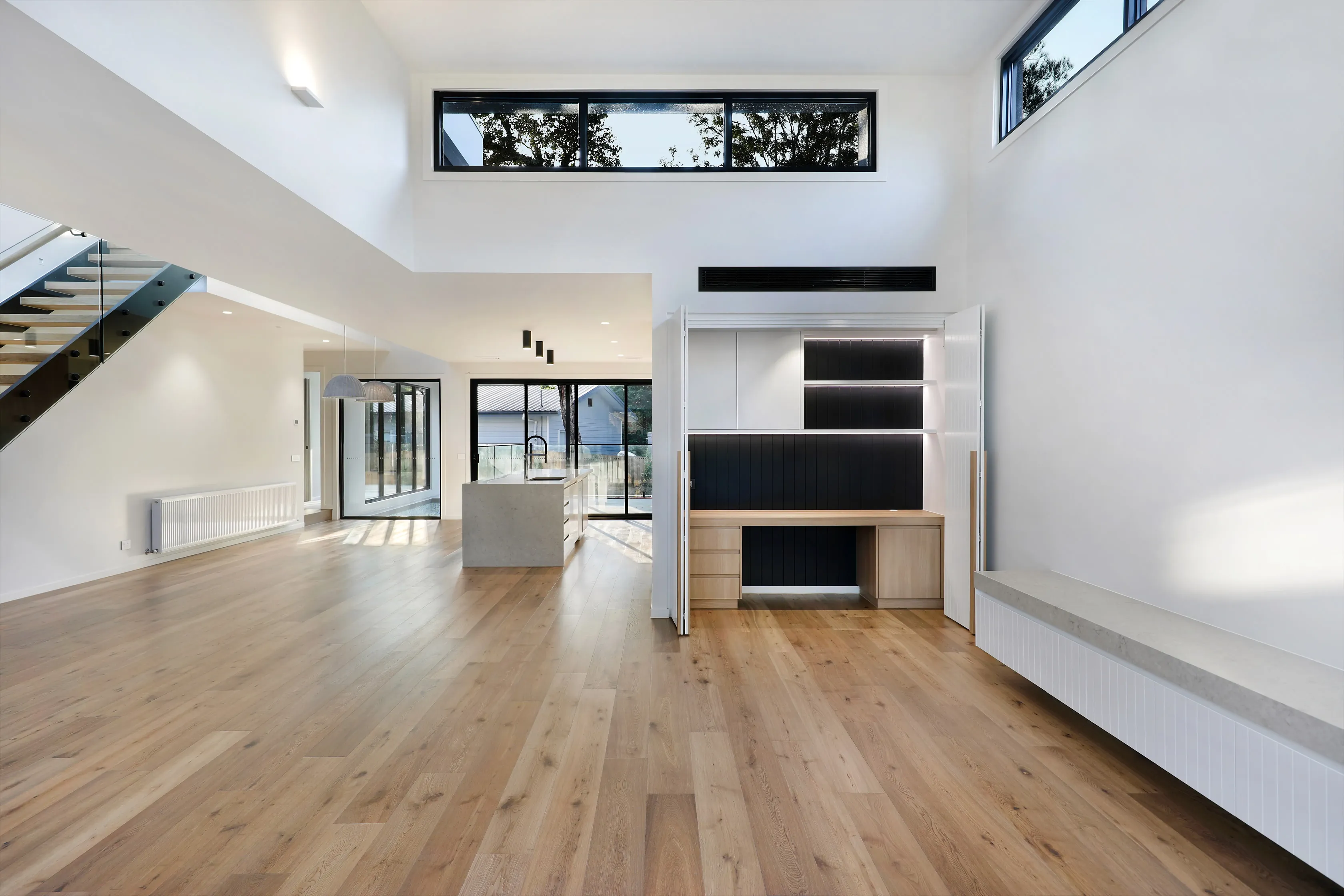 Double-height open living area with oak floors, a stair with glass balustrade, clerestory windows, a built-in oak study nook with black shelving and a stone window bench