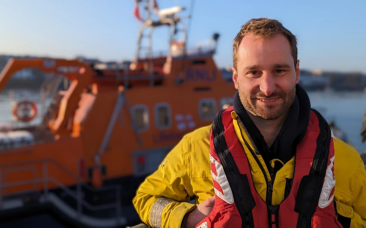 A person wearing a yellow waterproof jacket and a red and black life jacket stands on a dock with an orange rescue boat in the background. The boat is equipped with antennas and safety markings, and the scene is set on calm water with a clear sky, suggesting a maritime or lifeboat crew environment.