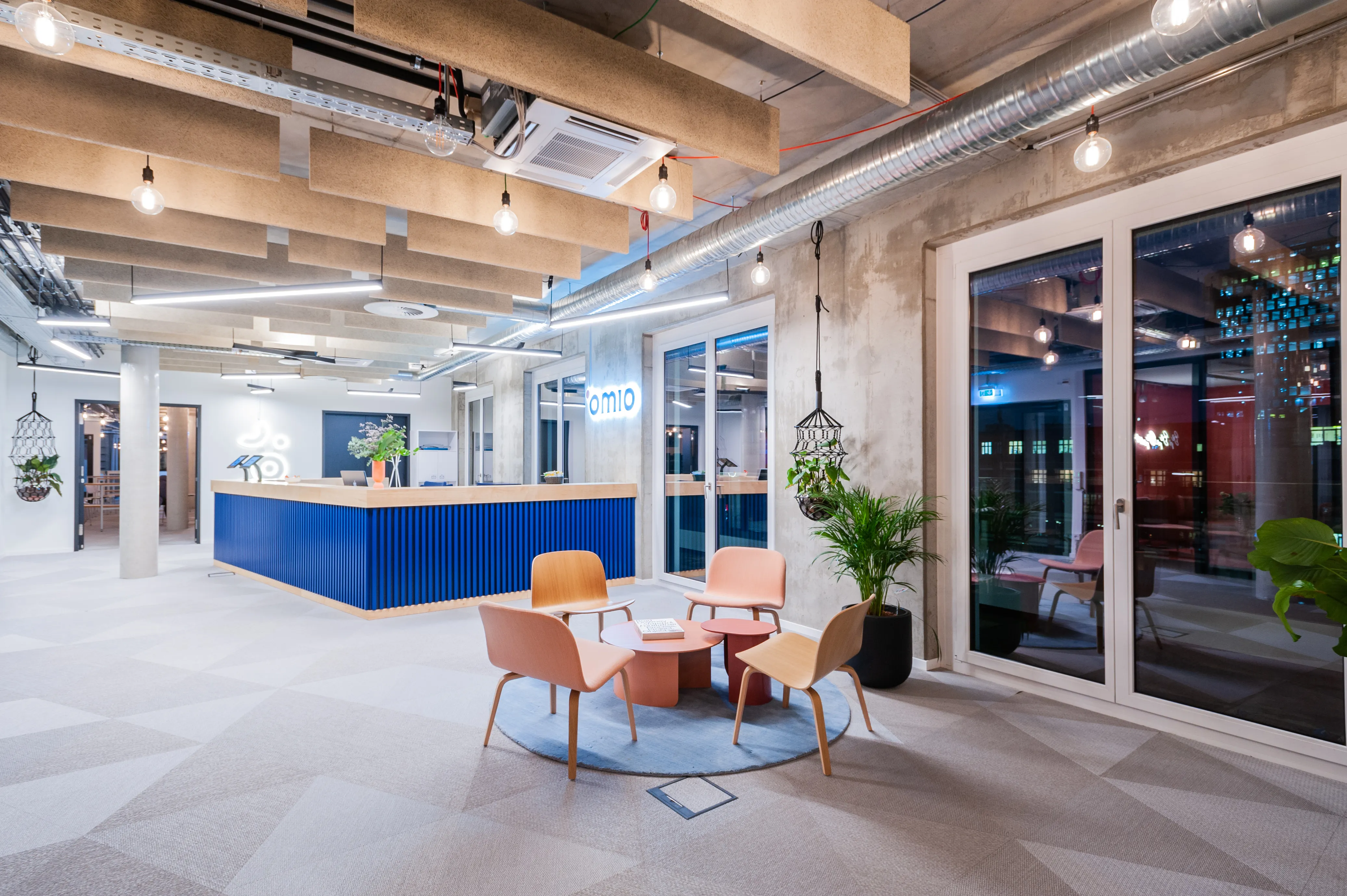 Entrance lounge at OMIO with the blue slatted reception desk in the background, terracotta and pink wooden lounge chairs around a small table on a round blue rug, and hanging glass pendants.