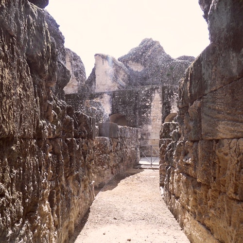 Pathway between ancient stone ruins with weathered, crumbling walls leading to a partially enclosed area. Bright, sunlit scene.