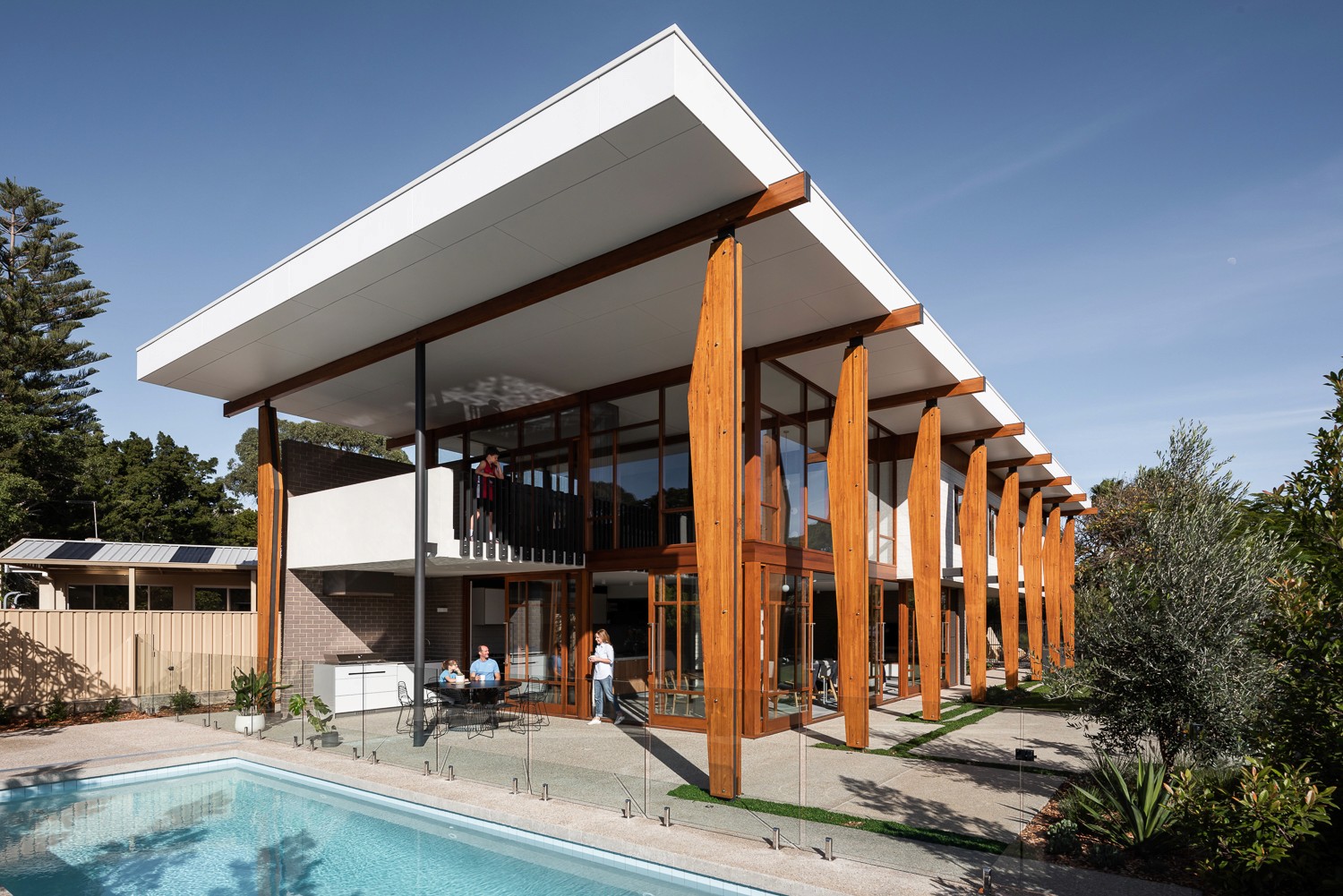 A modern house with large windows and a flat roof, surrounded by greenery against a clear blue sky.