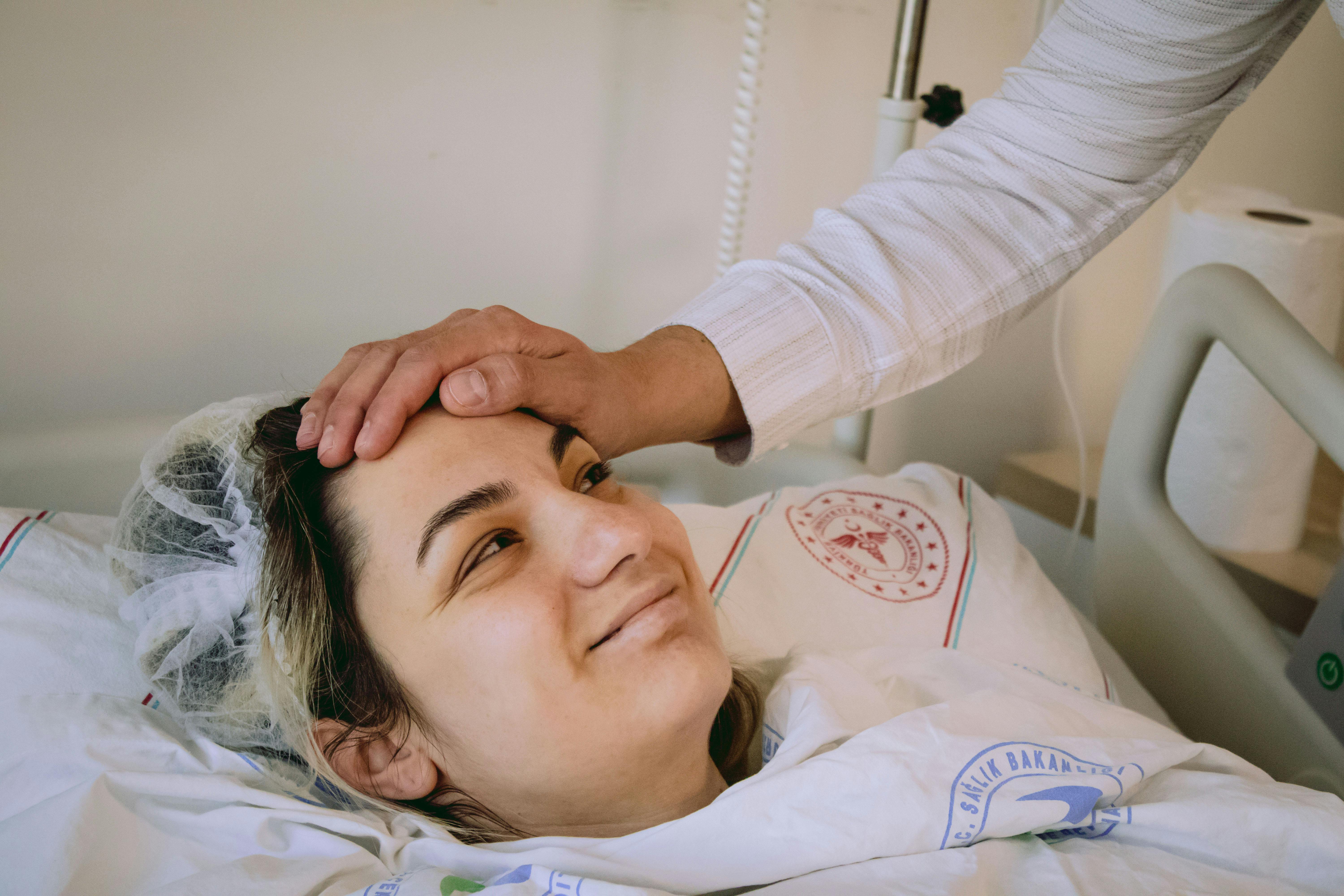 A woman lies on a hospital bed, she is wearing a hair net and smiling up at a person standing over her with their hand on her forehead.