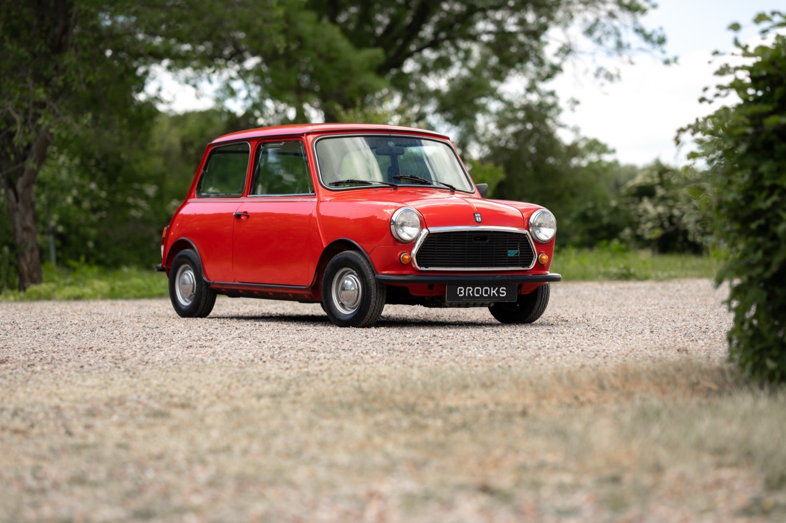 A red vintage car parked on a gravel road surrounded by greenery and a few trees in the background.