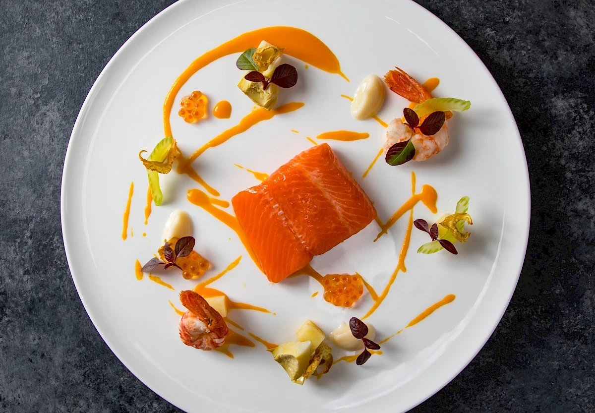 Gourmet-Gericht mit einem perfekt gegarten Lachsfilet, garniert mit Kaviar, Garnelen und kunstvollen Saucen-Akzenten auf einem weißen Teller.