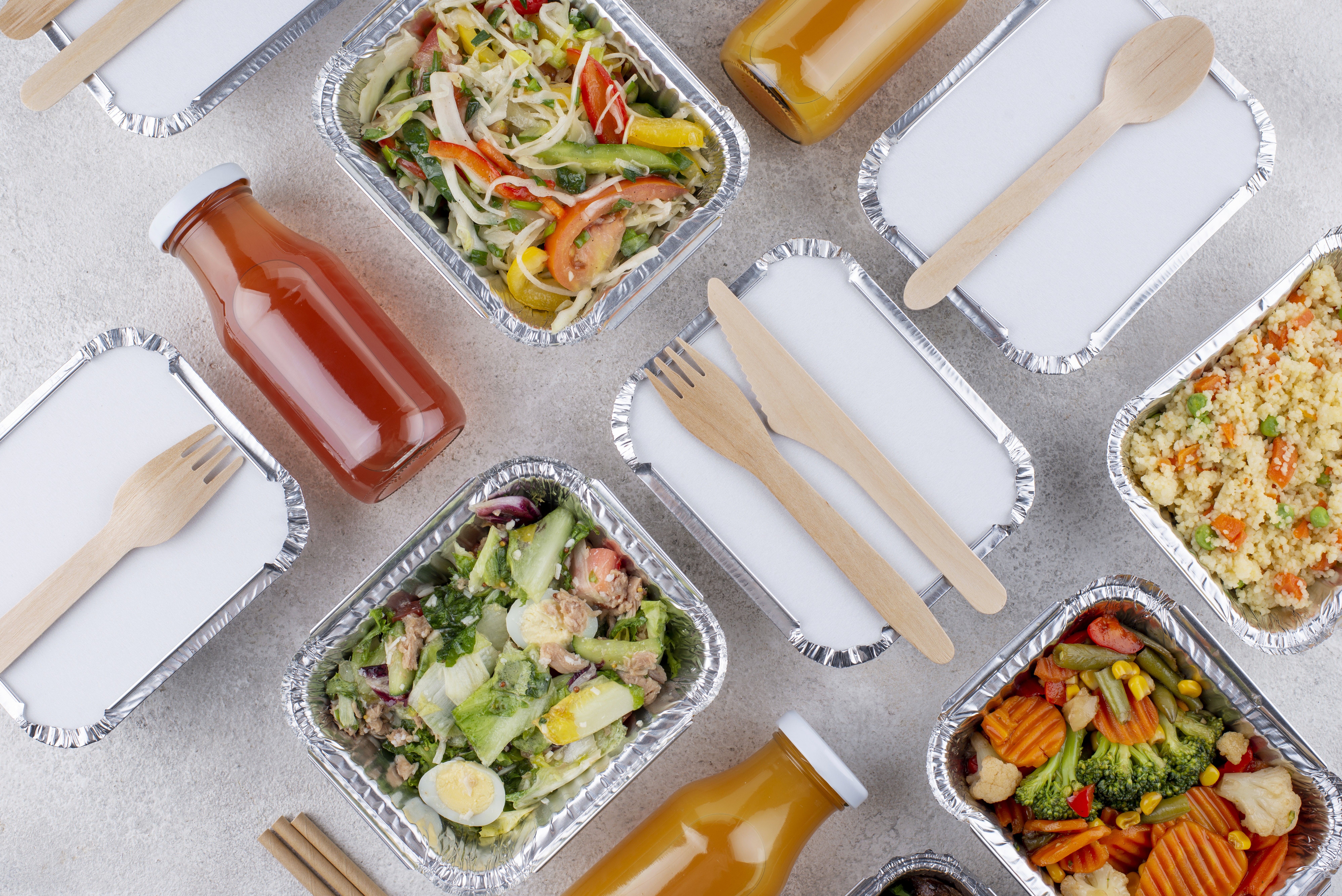 Na zdjęciu widać starannie ułożony zestaw gotowych posiłków w aluminiowych pojemnikach. Wypełniają je różnorodne dania — od sałatek z jajkiem, pomidorami i ogórkiem, po mieszanki warzywne z brokułem, marchewką i papryką oraz lekkie dania z kaszą i warzywami. Obok stoją butelki z sokami w dwóch kolorach, pomarańczowym i czerwonym, sugerujące różne smaki. Na części pojemników położono drewniane sztućce, co podkreśla, że posiłki są gotowe do zabrania i zjedzenia.