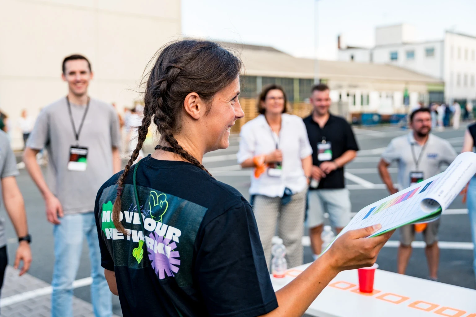 Eine junge Frau mit Zöpfen hat eine Mappe in der Hand, vor ihr steht eine Gruppe an Menschen, sie befinden sich in einer Workshop-Situation.
