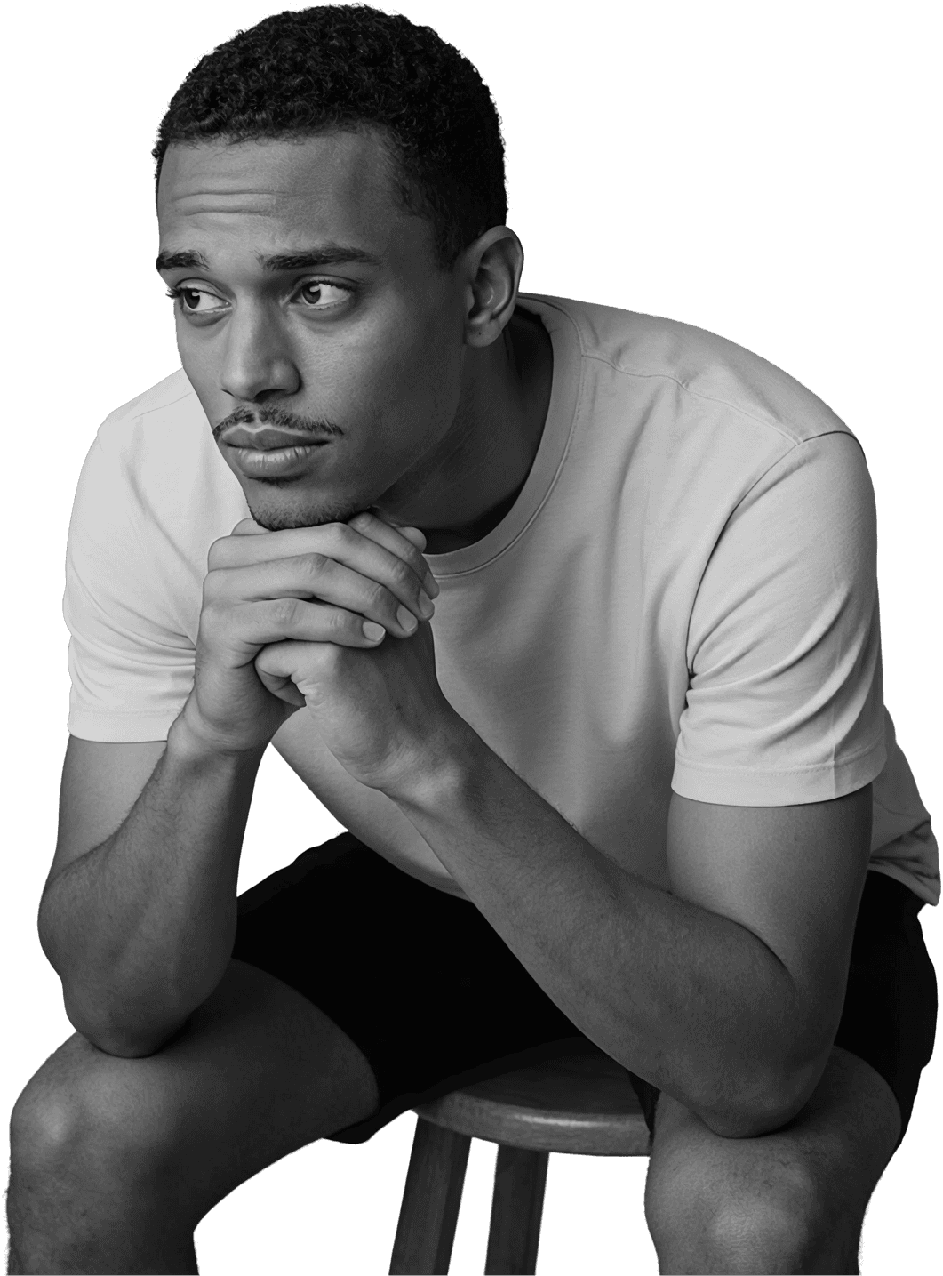 Pensive young man sitting on a stool with hands clasped under his chin, looking away in a dark studio setting