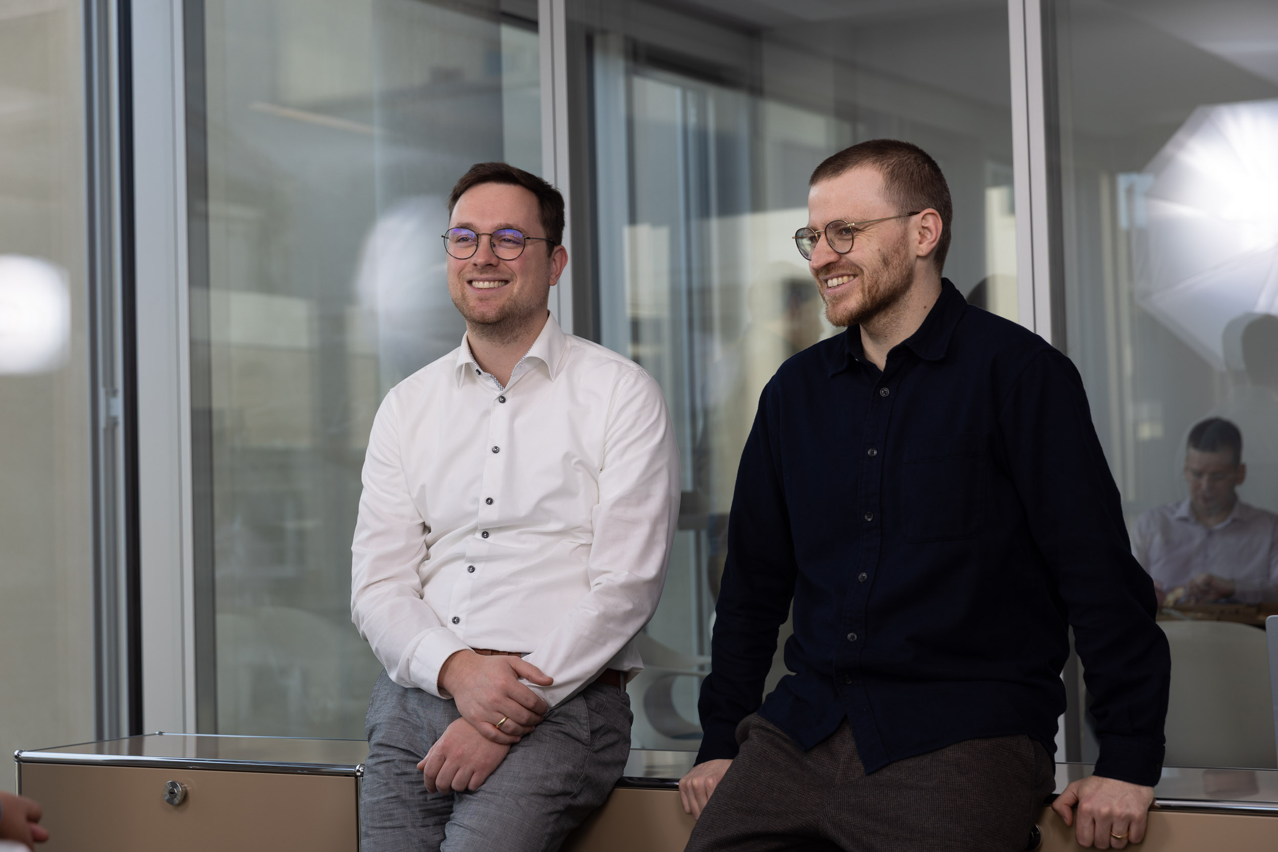 Zwei Mitglieder des Indicium Teams sitzen im Büro vor einer Fensterfront und lächeln in die Kamera