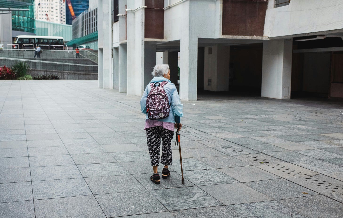 Older woman walks away from camera with cane, alone in a concrete urban or suburban cooridor.