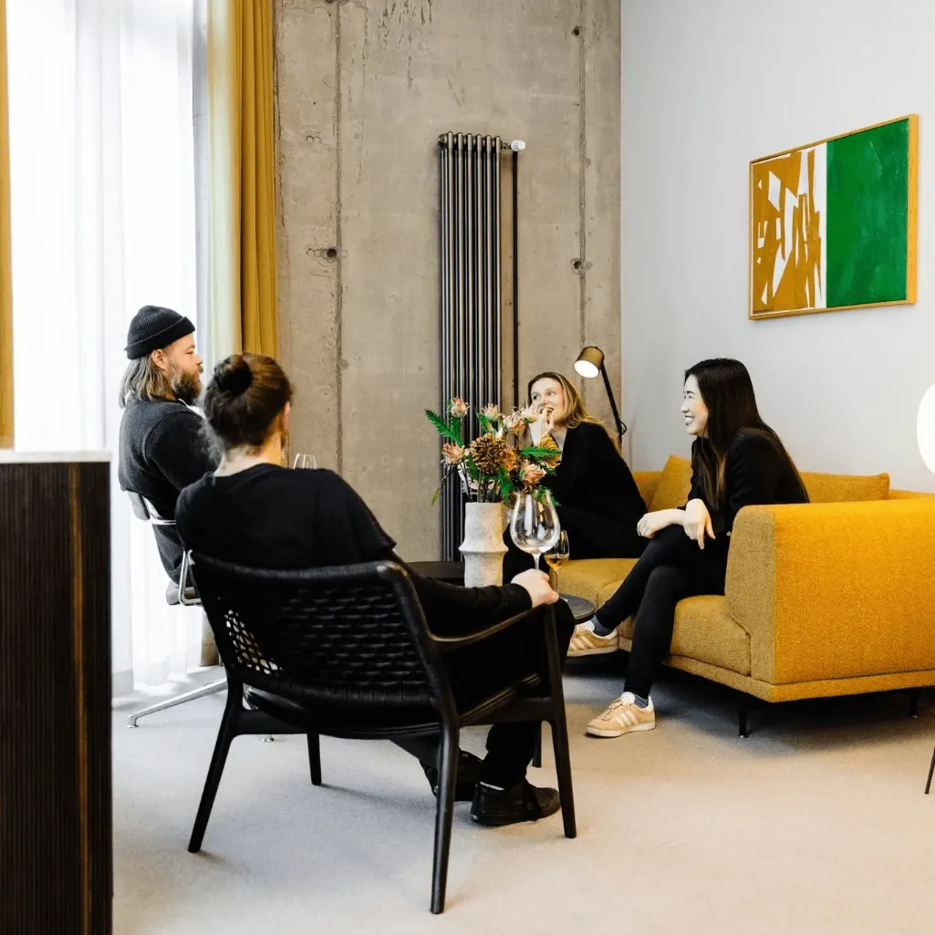 Four people gathered in a bright lounge, talking around a small table with flowers and wine glasses beside a mustard-yellow sofa and framed abstract artwork.