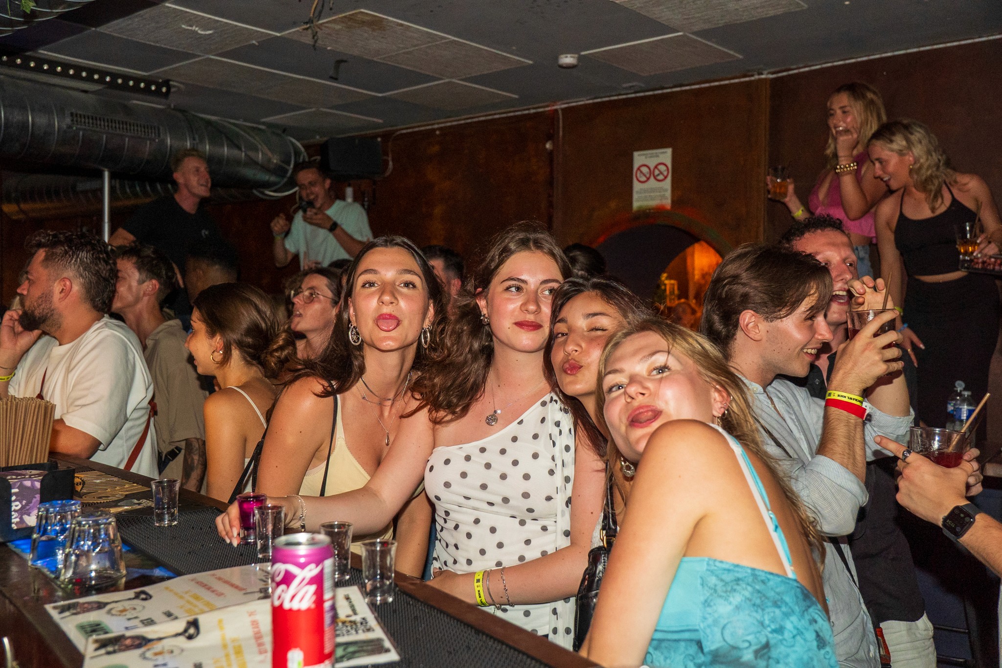 Group of friends posing and laughing at the bar during a bar crawl in Nice with drinks on the counter capturing social bonding playful energy and the vibrant nightlife atmosphere of a crowded party venue on the French Riviera
