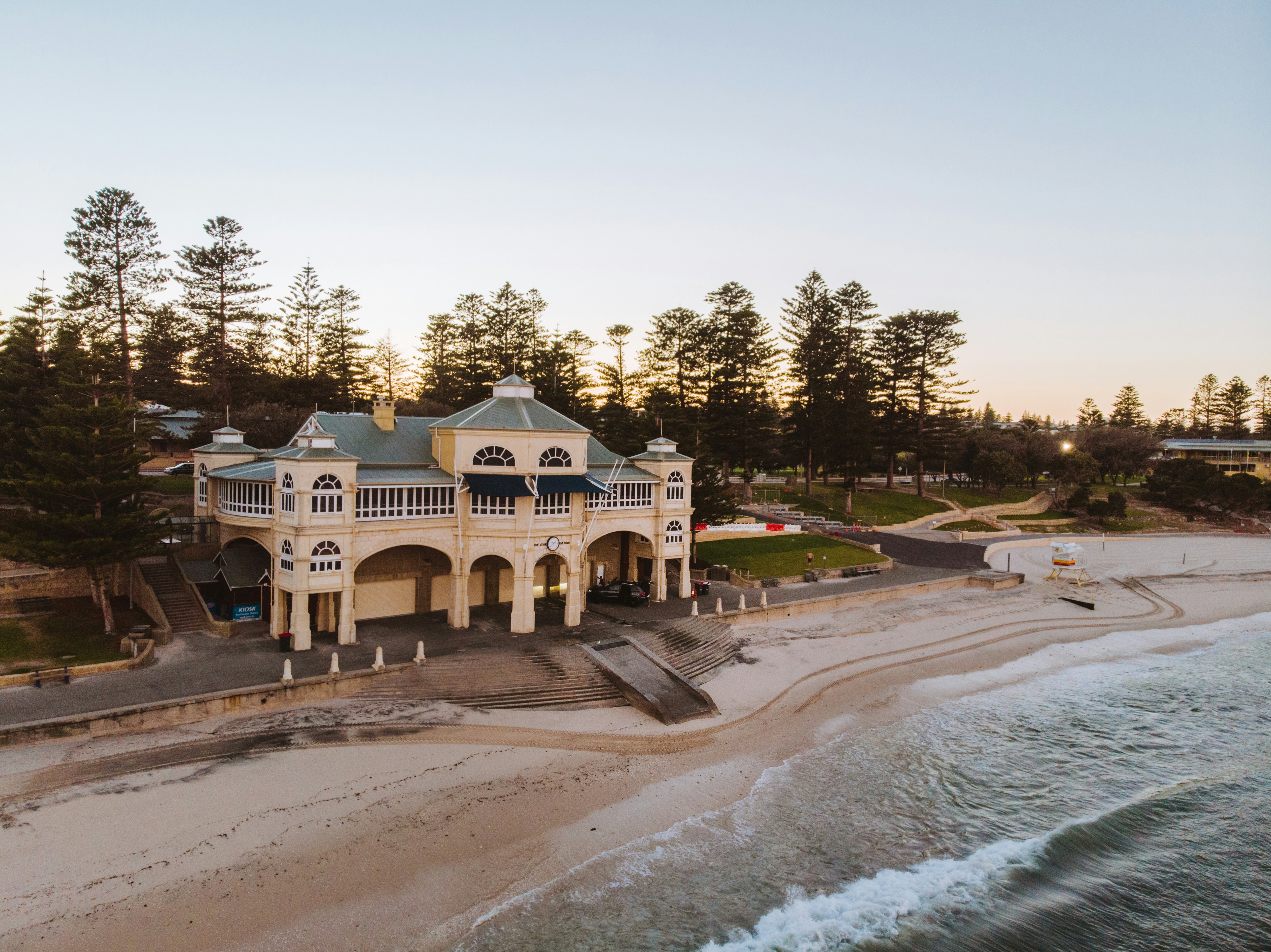 Coastal mansion near the shore at sunset, surrounded by trees and gentle waves lapping at the beach.