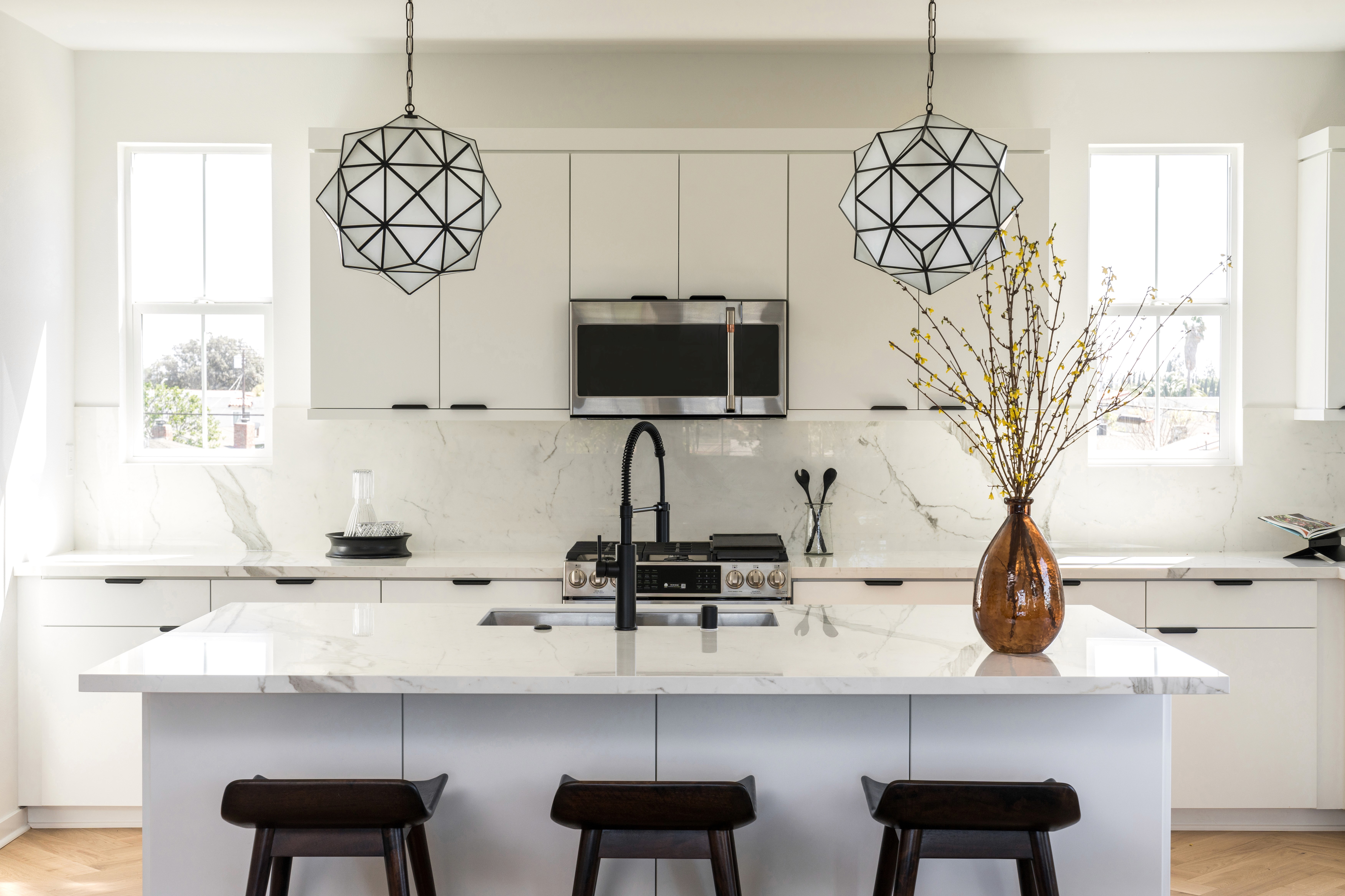 Dining area with black boucle dining room chairs, modern, glass top dining table, black pedestal bowl and black and white striped vase; kitchen in background with flat white cabinets, black hardware, white marbled countertops