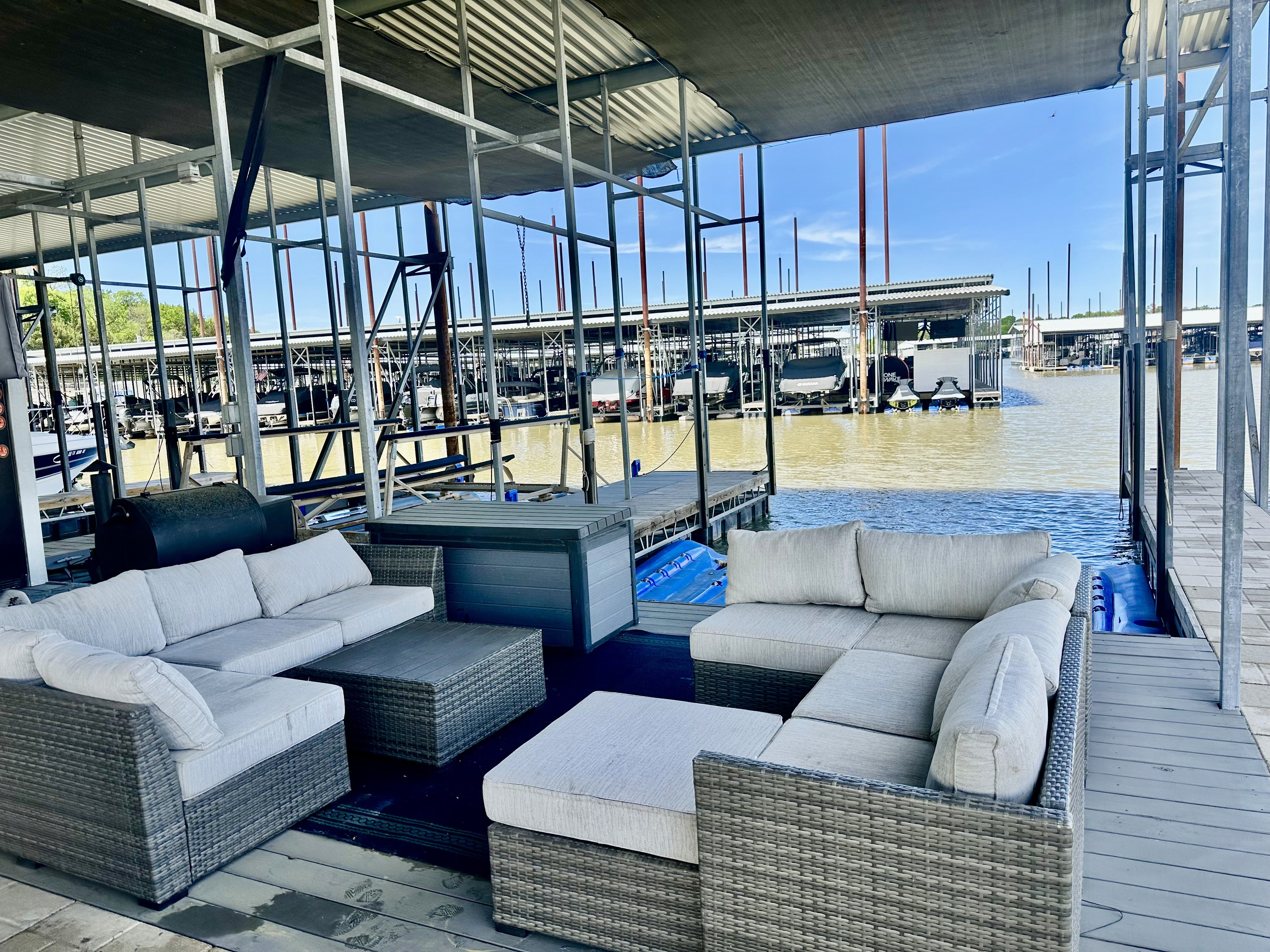 Outdoor seating area on a dock with gray wicker sofas and white cushions overlooks a marina filled with boats under clear blue skies.