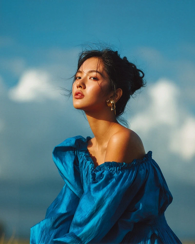 A woman in a vibrant blue blouse poses against a serene sky, her gold earrings catching the light, creating a striking and elegant portrait.