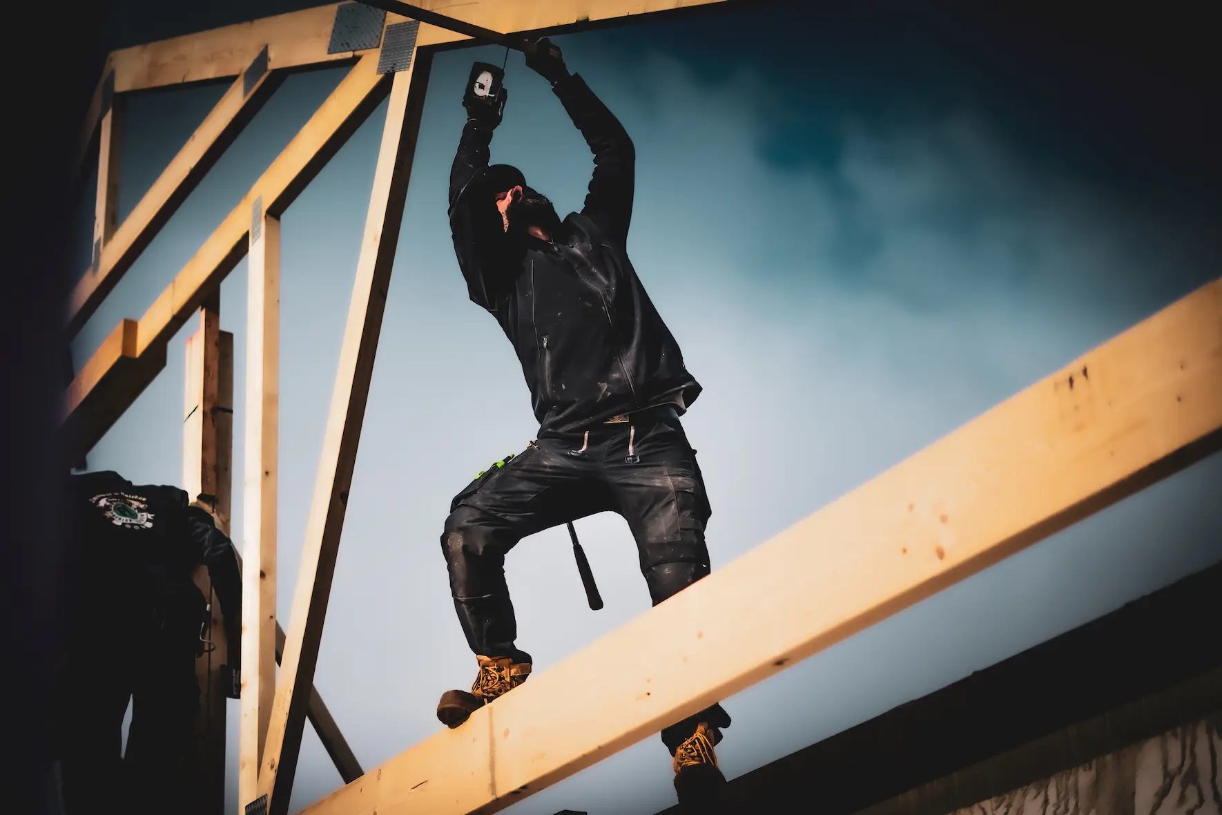 Ein Bauarbeiter in Schutzkleidung balanciert geschickt auf Holzbalken vor dem Hintergrund eines bewölkten Himmels und stellt dabei sein Fachkönnen in den Bereichen Dacheindeckung und Montage von Tragwerken unter Beweis.