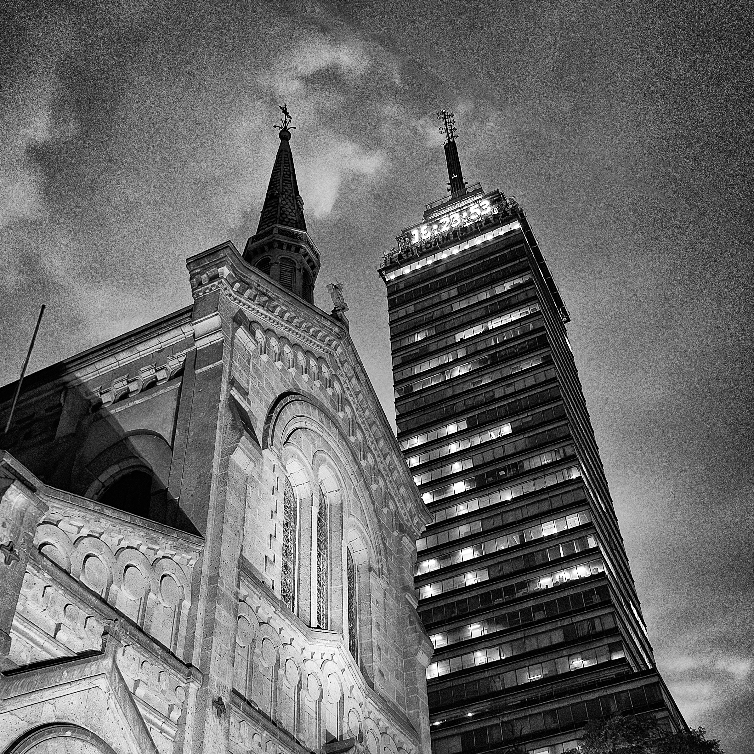 Black and white photo of a church facade beside the Torre Latinoamericana in Mexico City, both illuminated under a dramatic evening sky.