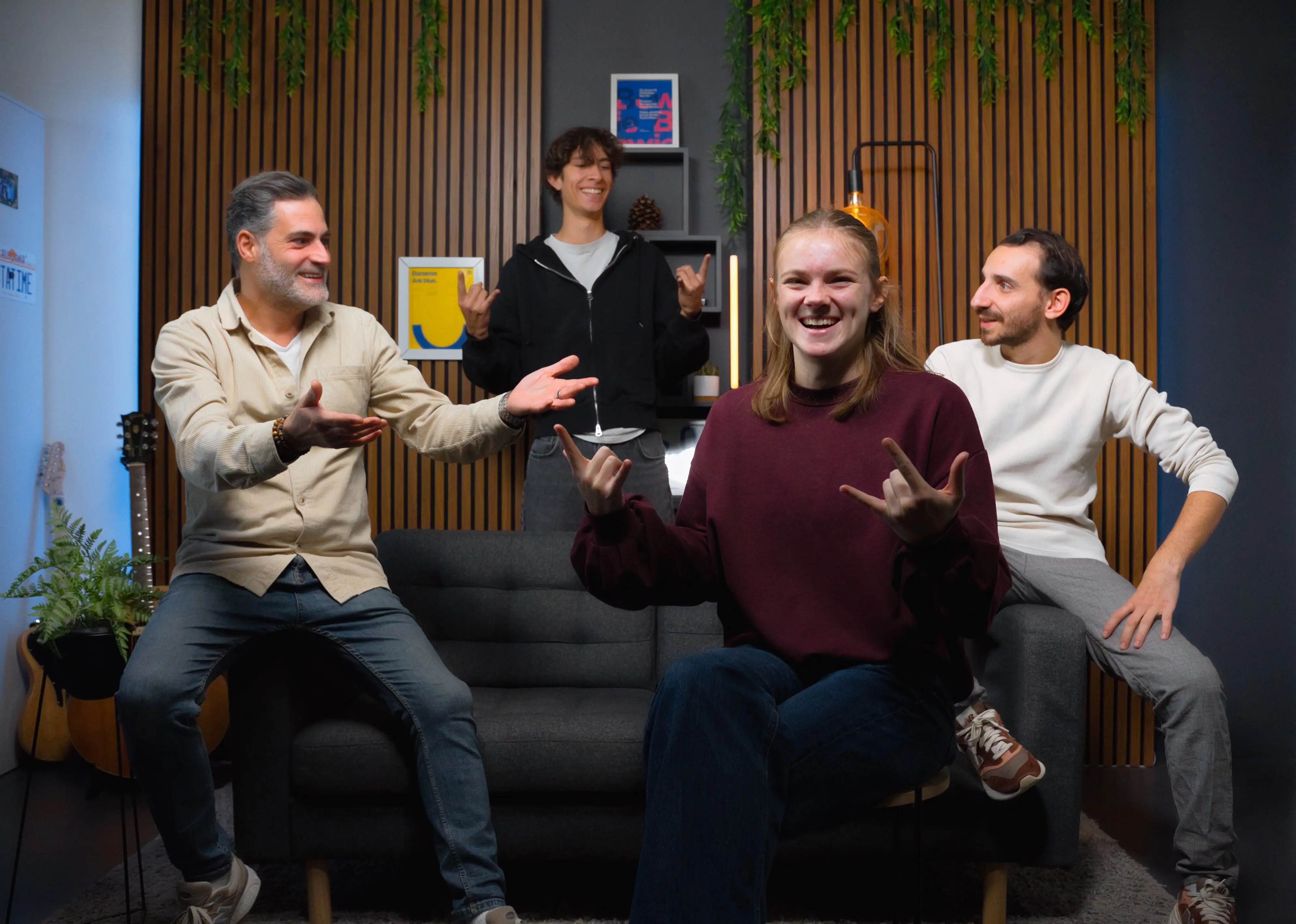 Four people are gathered in a modern living room setting featuring a black sofa, wood-paneled walls, and decorative plants, with some engaging in conversation and others gesturing playfully.