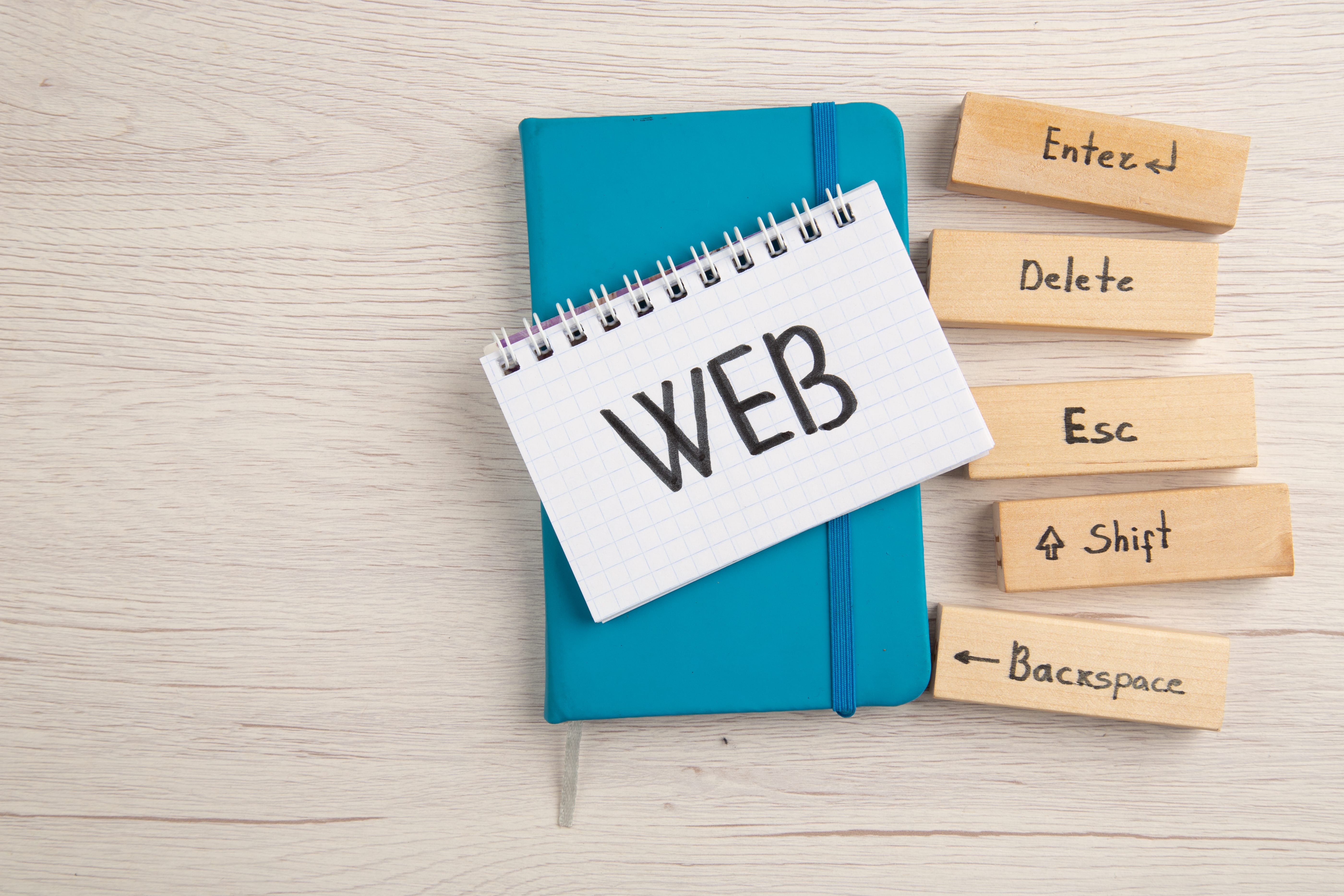 Top view of notepad with 'WEB' text on blue notebook next to wooden keyboard key blocks labeled Enter, Delete, Esc, Shift, and Backspace on light wooden background