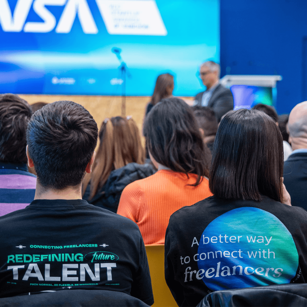 Two people at a wooden desk with a laptop and coffee, chatting and laughing by a large window