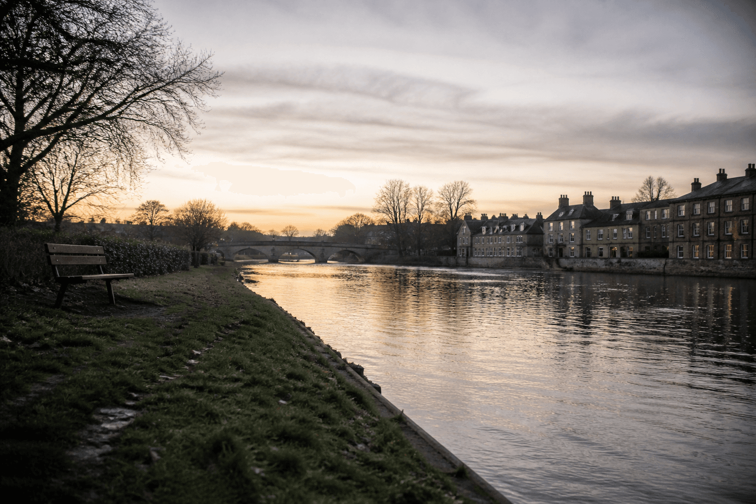 Quiet riverside scene along the River Cam in Cambridge at early evening with a bench, calm water, and soft natural light.