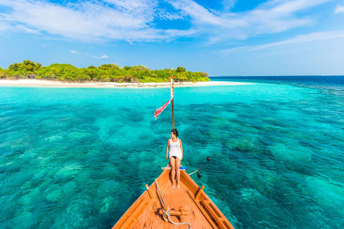 A person stands on the bow of a wooden boat in turquoise water facing a tropical island with green trees and white sand.