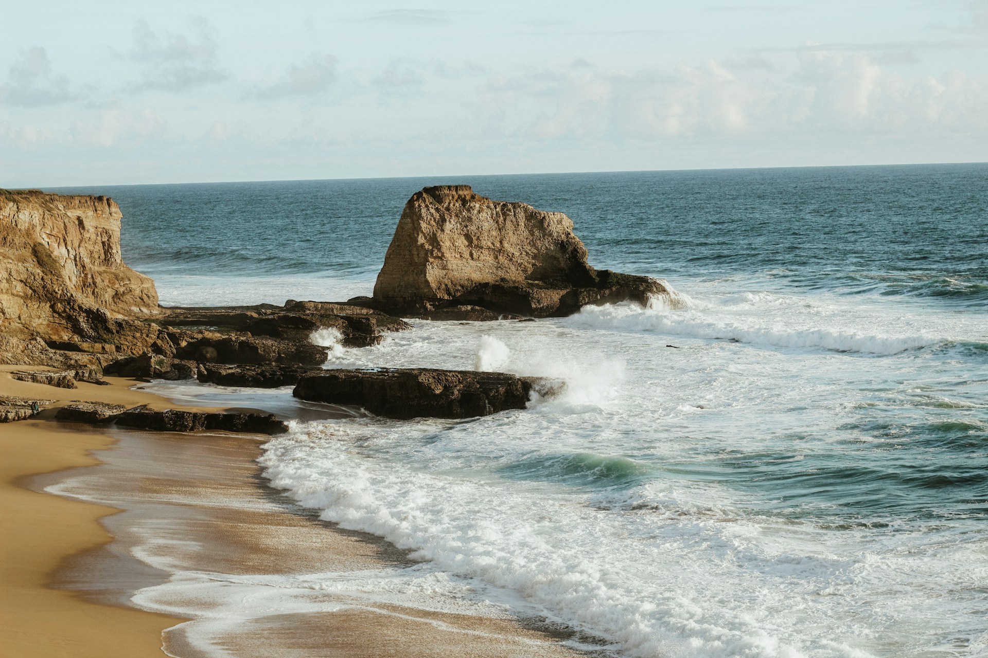 Rocky sea stacks and churning surf along a sandy coastline under a cloudy sky.