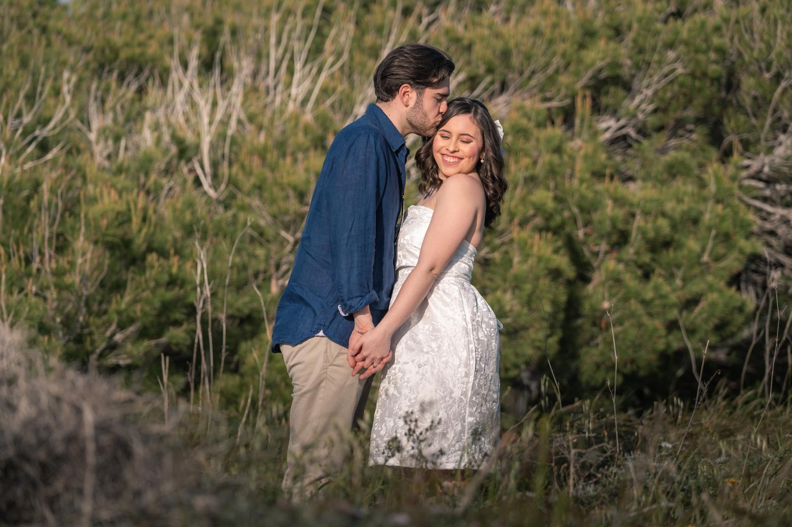 A couple holds hands, standing on a rocky beach under a large cave arch, gazing at the tranquil ocean during sunset. The sky is softly lit in pastel hues.