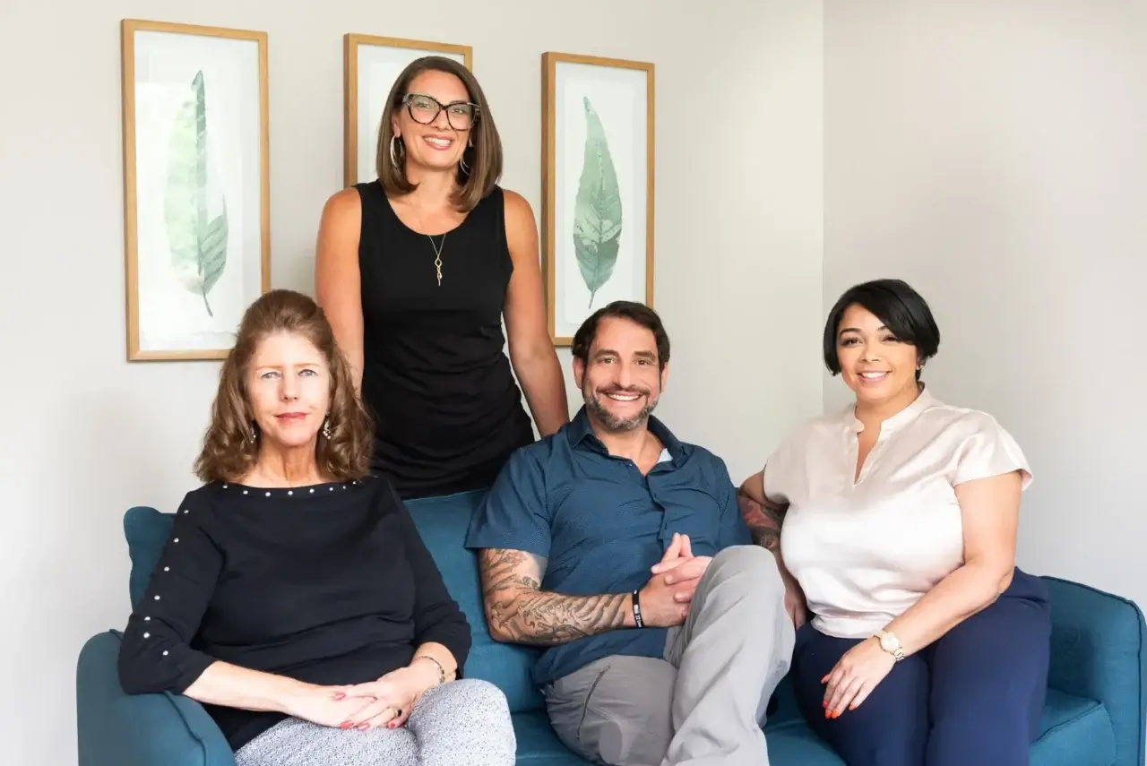 Professional team of four smiling on a blue couch in a modern office setting.