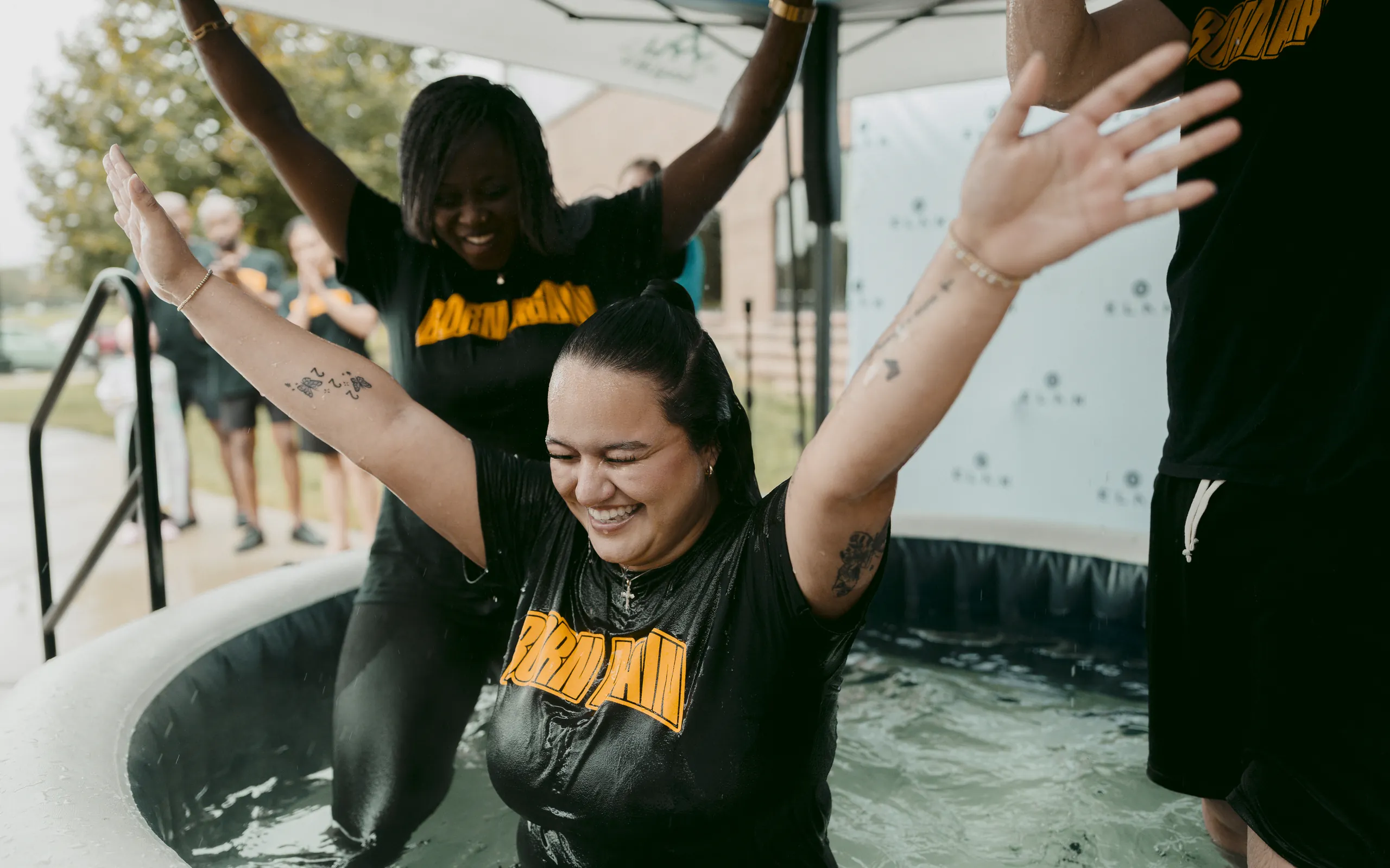Young woman raising her arms in joy at her baptism, celebrated by friends at Elan Church in Naperville, IL