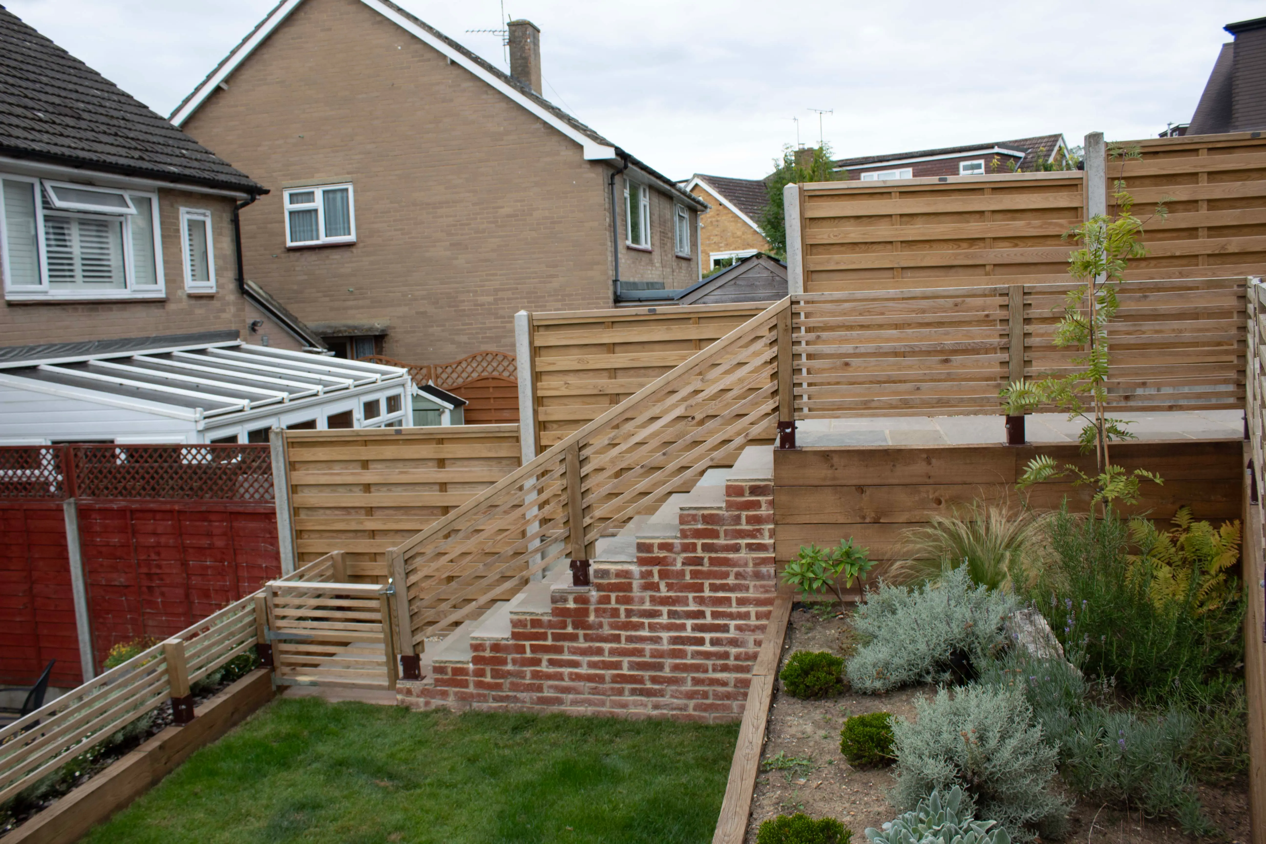 A garden area with wooden fences, paving stones, and green plants, bordered by neighboring houses.