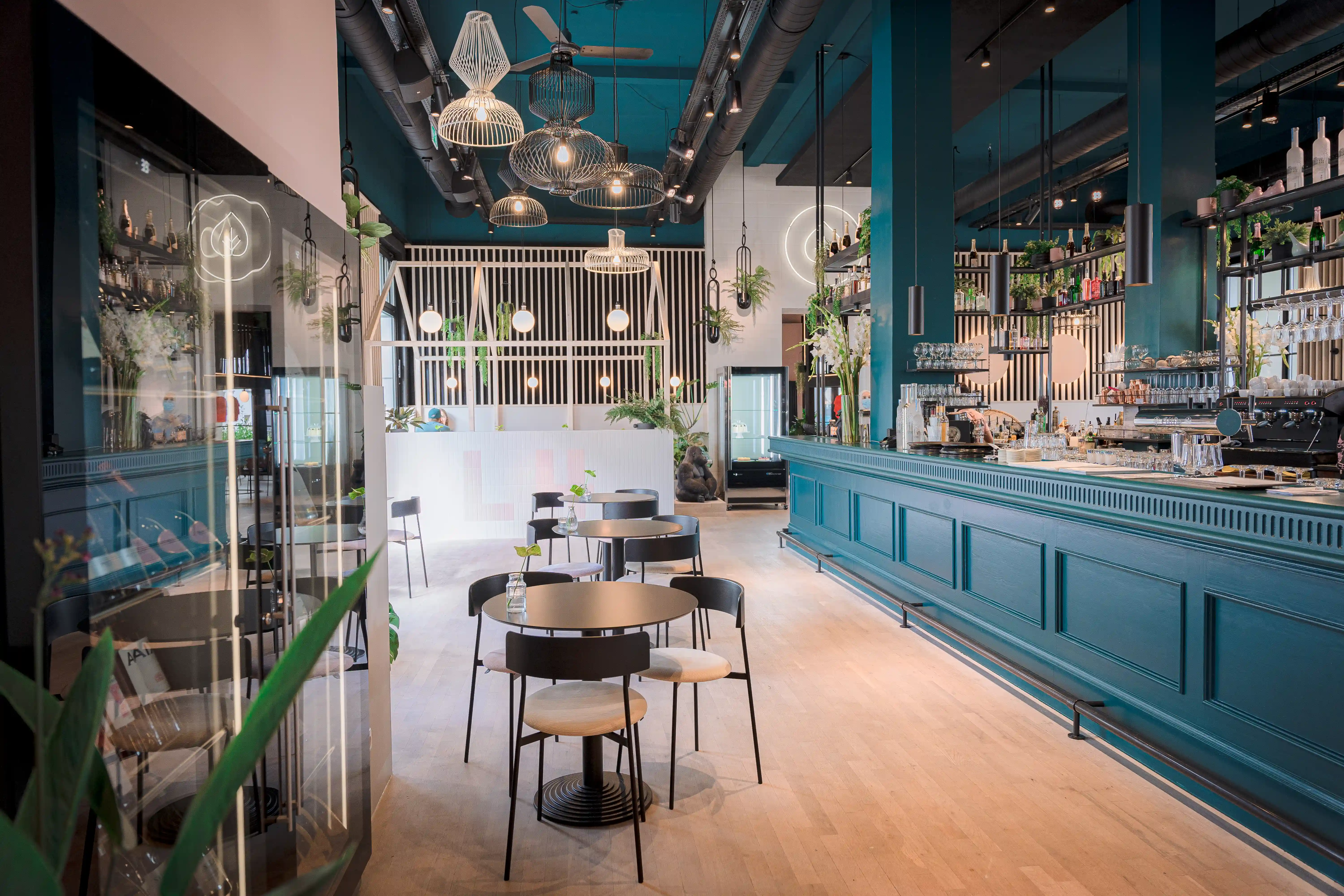 Long teal bar counter at LOKAL VIER with brass details, espresso machines and pastry display, black chairs at round tables and woven pendant lamps under a teal ceiling.
