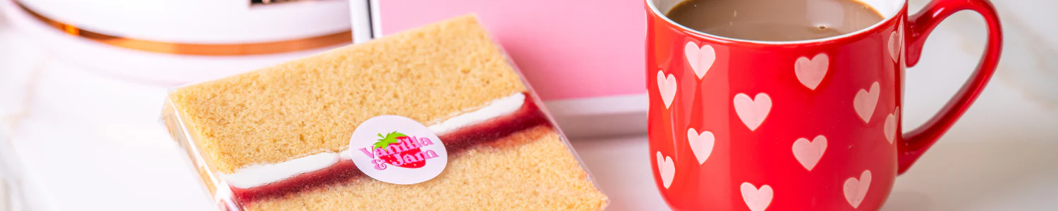 A vanilla cream-filled cake slice with a pink 'Vanilla & Jam' sticker, displayed next to a red coffee mug decorated with white hearts.