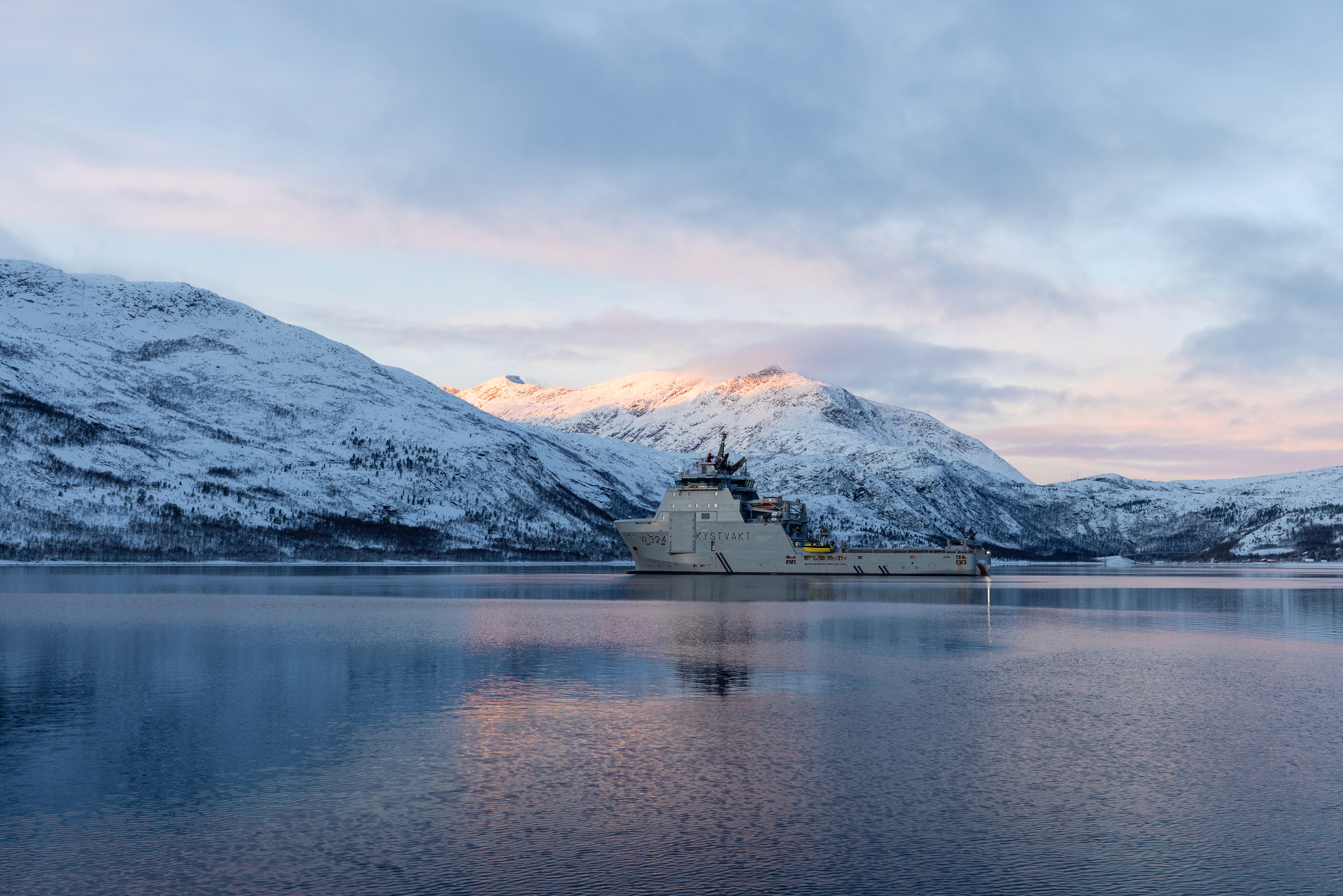 A serene winter landscape with snow-capped mountains reflecting in calm waters under a cloudy sky.