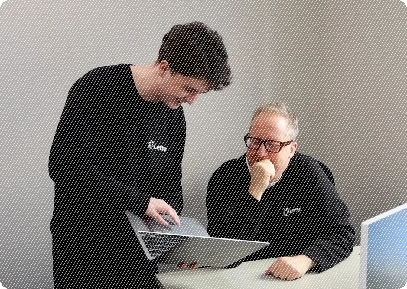 Two people in black attire are engaged in a collaborative discussion over an open laptop at a white desk, suggesting teamwork in a professional setting.