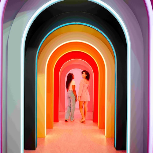 Due donne in piedi sotto archi illuminati multicolori in un corridoio, che si fronteggiano.
