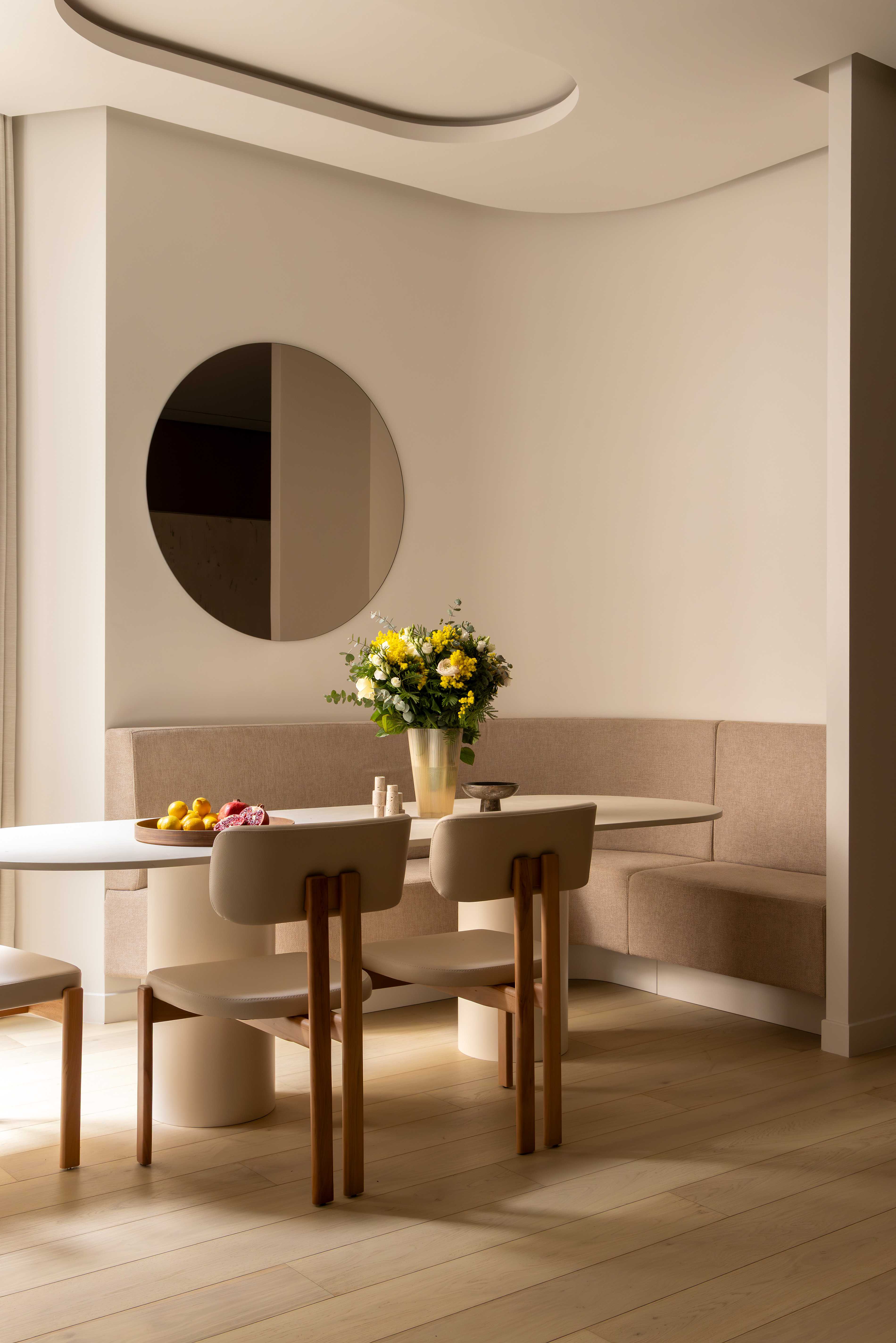 Espace repas avec banquette d'angle beige, chaises en bois et tissu, miroir rond, bouquet de fleurs et fruits