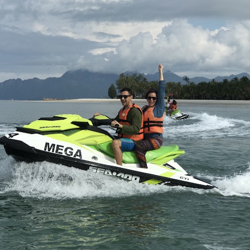 兩個人乘坐著檸檬綠色的水上摩托，穿著橙色救生衣，其中一個人舉起了一隻手，正在海灘和山脈附近的水面上行駛。