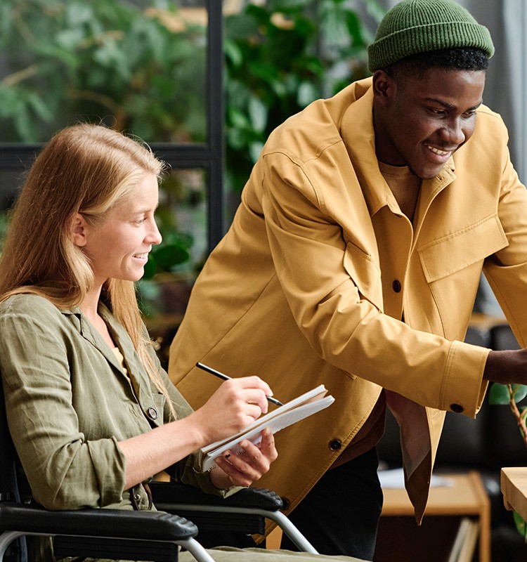  A woman in a wheelchair writing in a notebook while a man in a yellow jacket leans over to collaborate with her