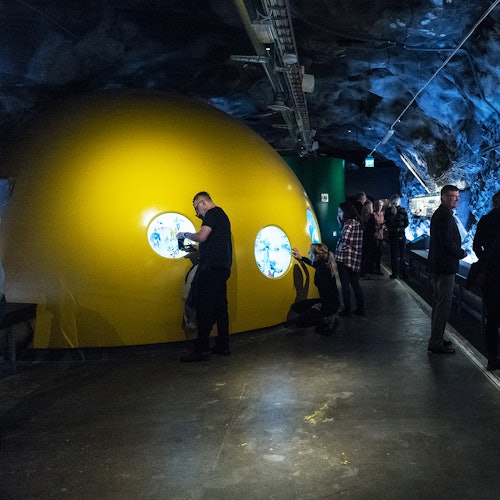 Personer som observerar utställningar genom fönster i en stor, gul, kupolformad struktur inne i ett svagt belyst tunnel-liknande rum.