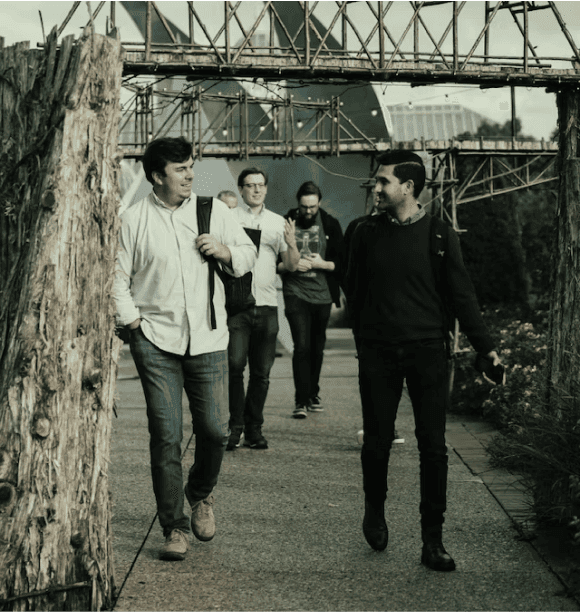 Four men walk together outdoors on a pathway, with two in front having a conversation and two walking behind them. A wooden archway and plants are visible along the path.
