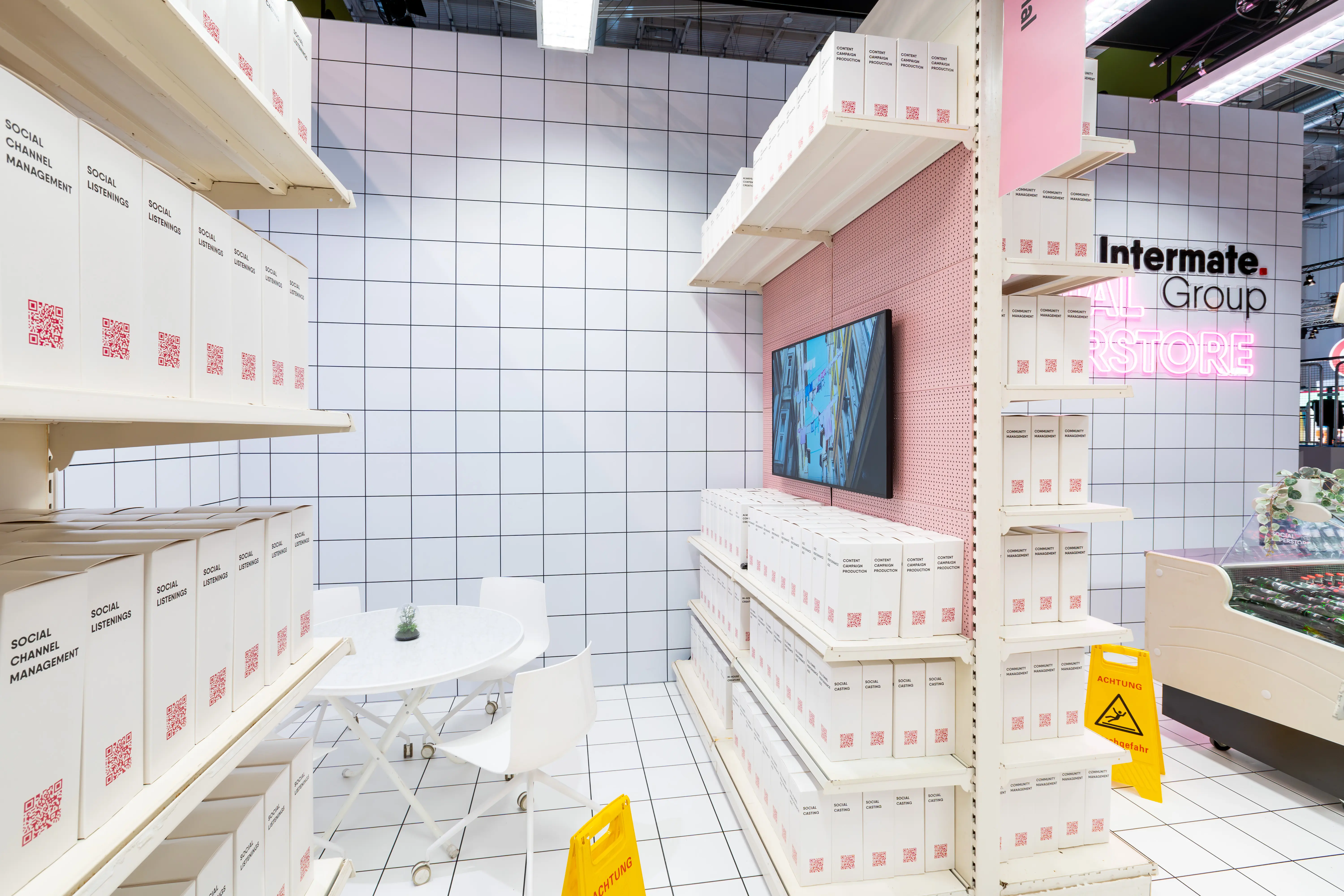 Interior of the Intermate Social Superstore booth with service shelves, screen on pink pegboard and white bistro table.