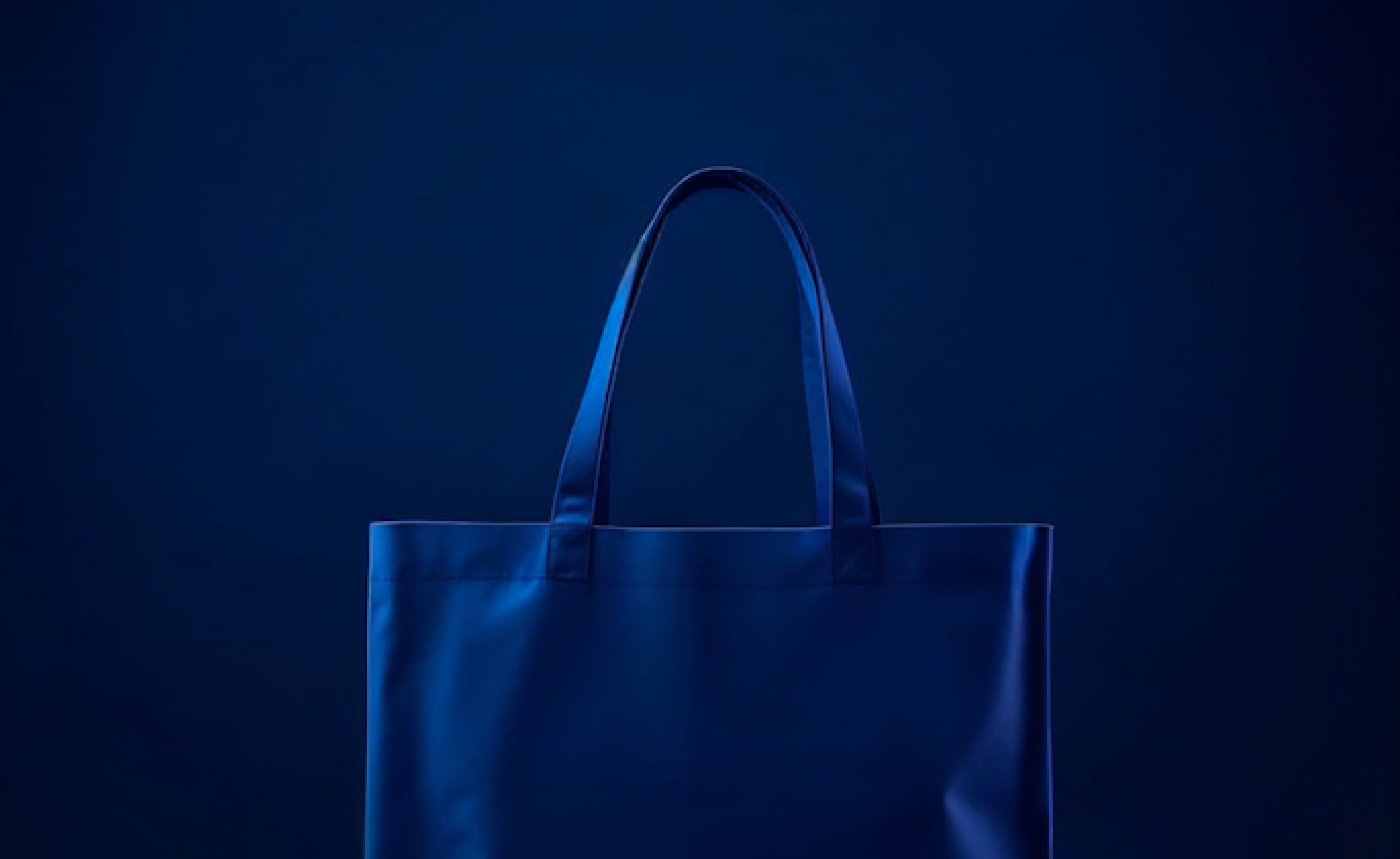 Dark blue tote bag with two handles on a matching blue background, lit to show texture.