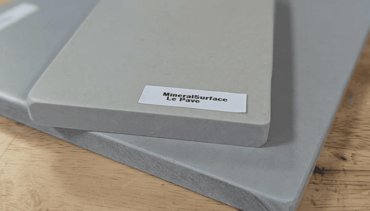 Trois dalles empilées de matériau composite gris clair portant l'étiquette MineralSurface Le Pave, posées sur un plan de travail en bois, montrant les faces lisse et brute du matériau