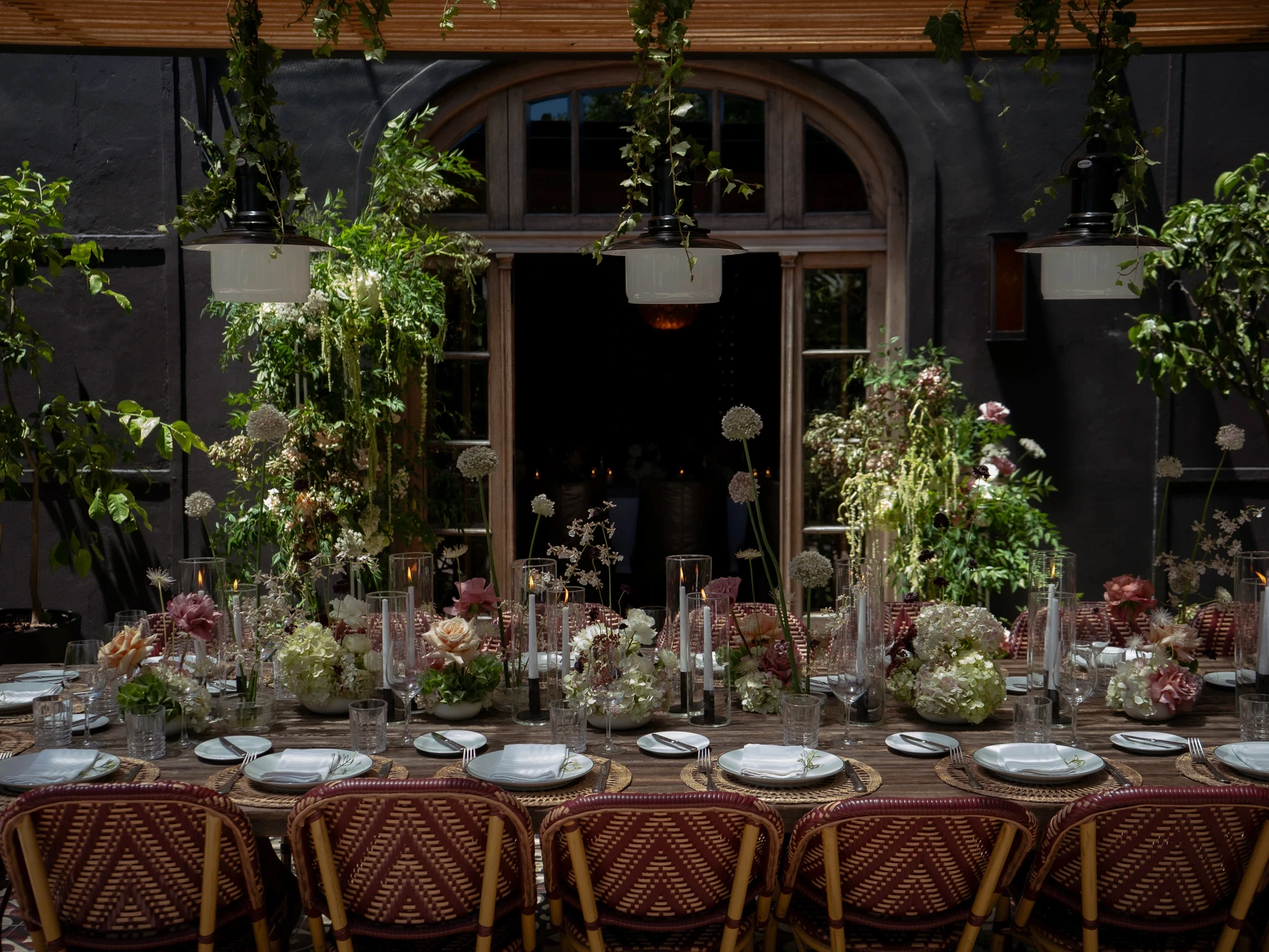 Mesa larga de madera en terraza con sillas de mimbre, arreglos florales bajos y lámparas colgantes blancas.