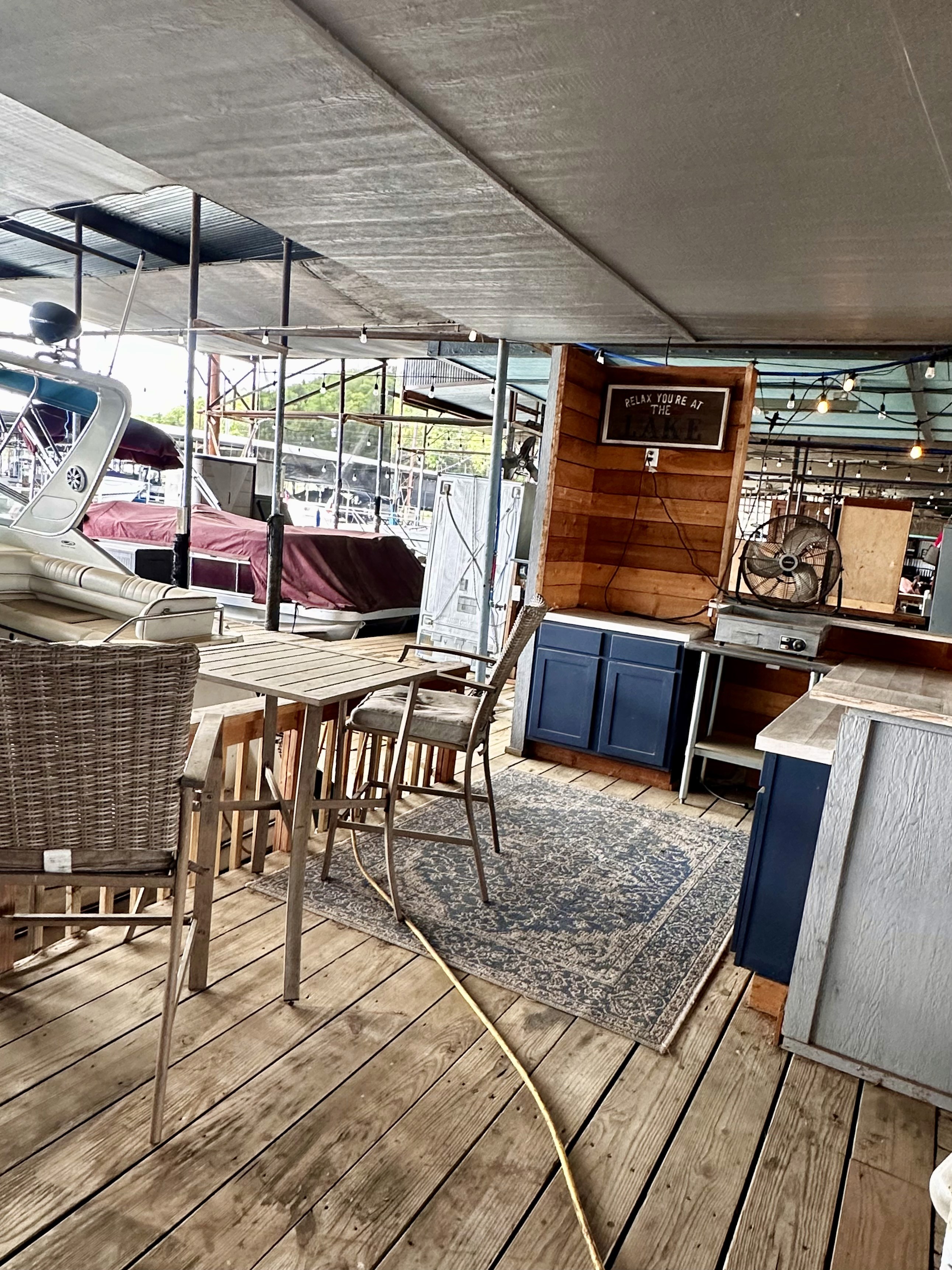 A spacious outdoor patio area features wooden flooring, stylish wicker seating around a wooden table, and a cozy rug, all set against a backdrop of boats in a marina under a covered canopy.