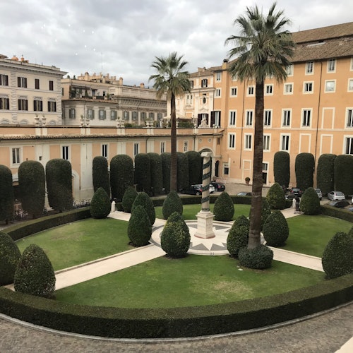 산책로, 다듬어진 울타리, 야자 나무, 중앙 조각상이 있는 잘 가꾼 안뜰 정원, 그리고 역사적인 건물들로 둘러싸여 있습니다.