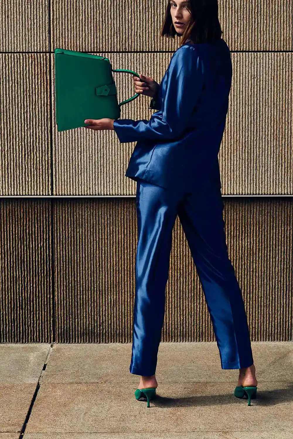 Model in a blue jumpsuit stands on textured pavement holding a green folding bag.