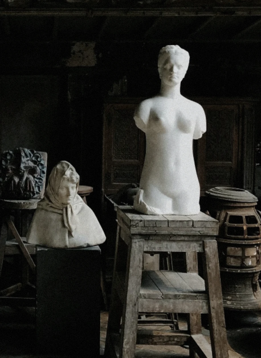 A dimly lit artist’s studio with classical marble sculptures — a veiled bust and a torso of a woman — resting on aged wooden stands, evoking timeless craftsmanship and quiet reverence.