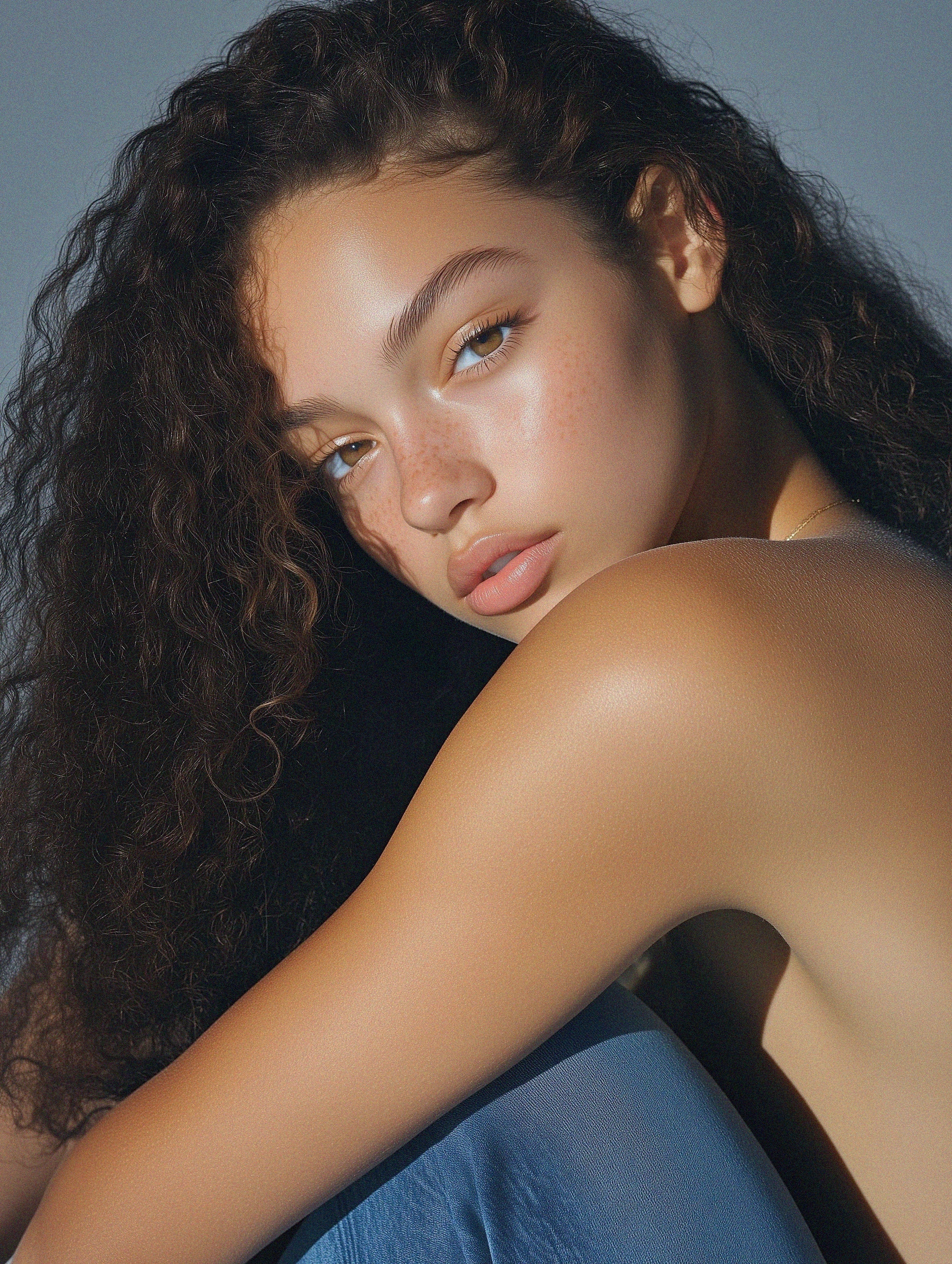 Portrait of a young woman with natural curly hair, freckles, and glowing skin, sitting in soft light against a neutral background.